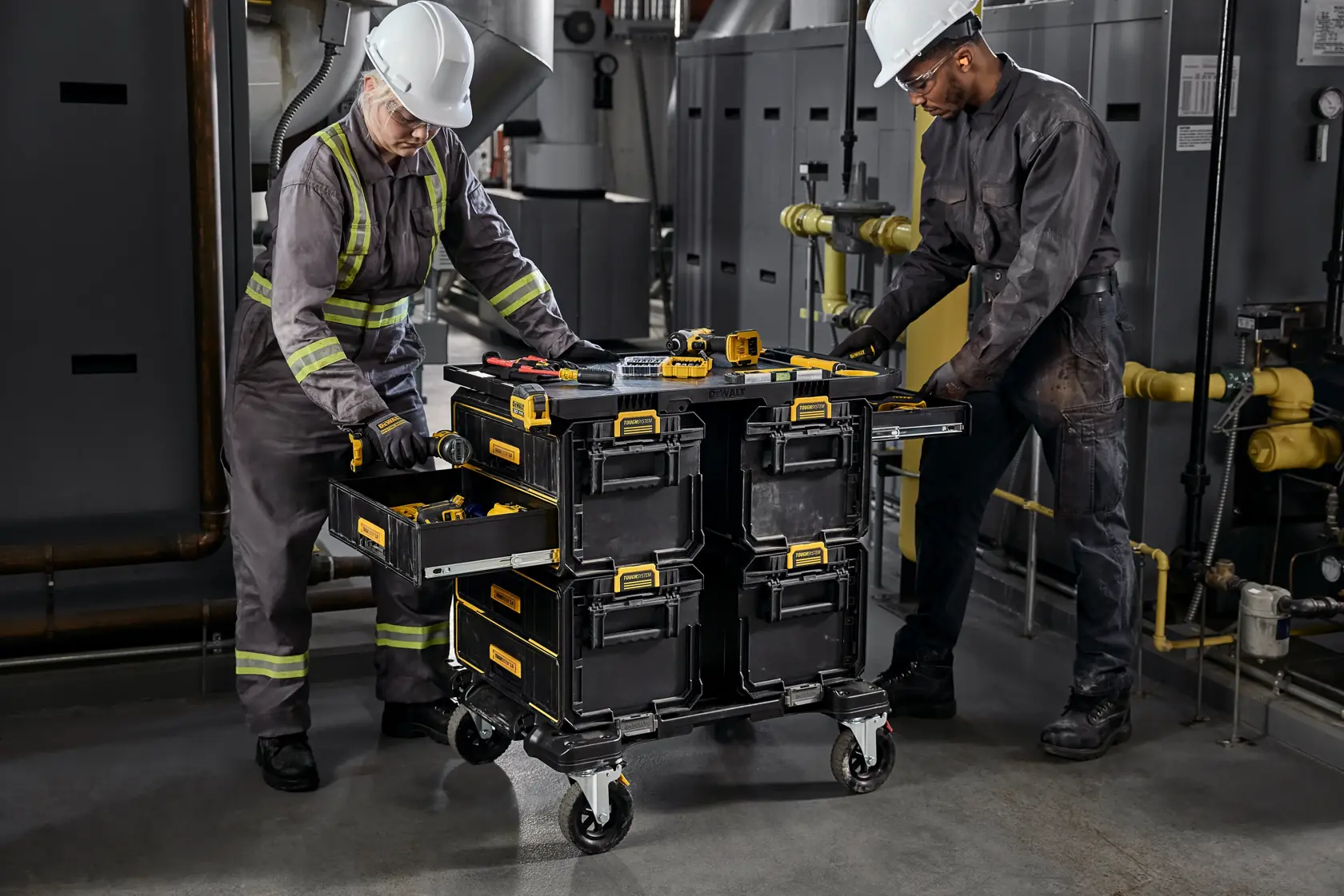 Two workers wearing safety helmets and reflective uniforms are organizing tools in an industrial setting. They use a DEWALT rolling tool storage cart with drawers and boxes, holding various tools on top and inside the compartments.