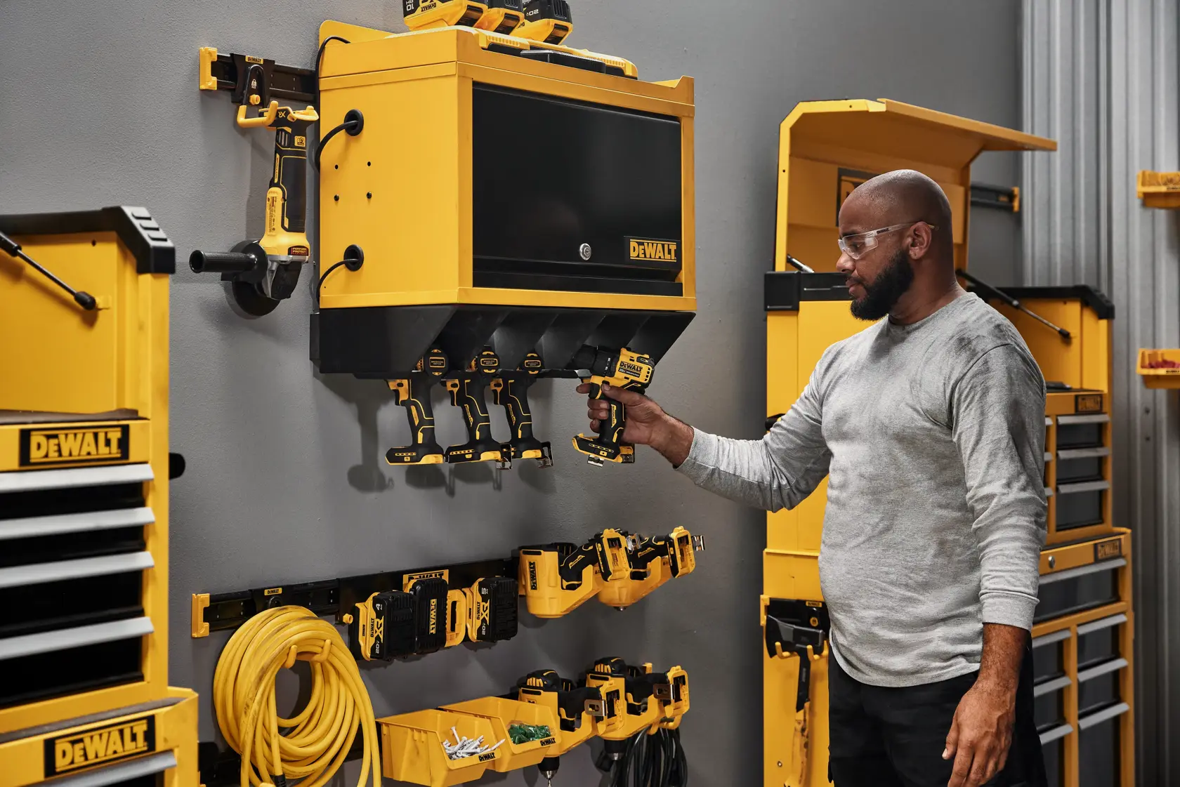 A person stands in a workshop next to a wall-mounted storage system featuring various DEWALT yellow and black power tools and accessories, including drills, chargers, and cabinets.