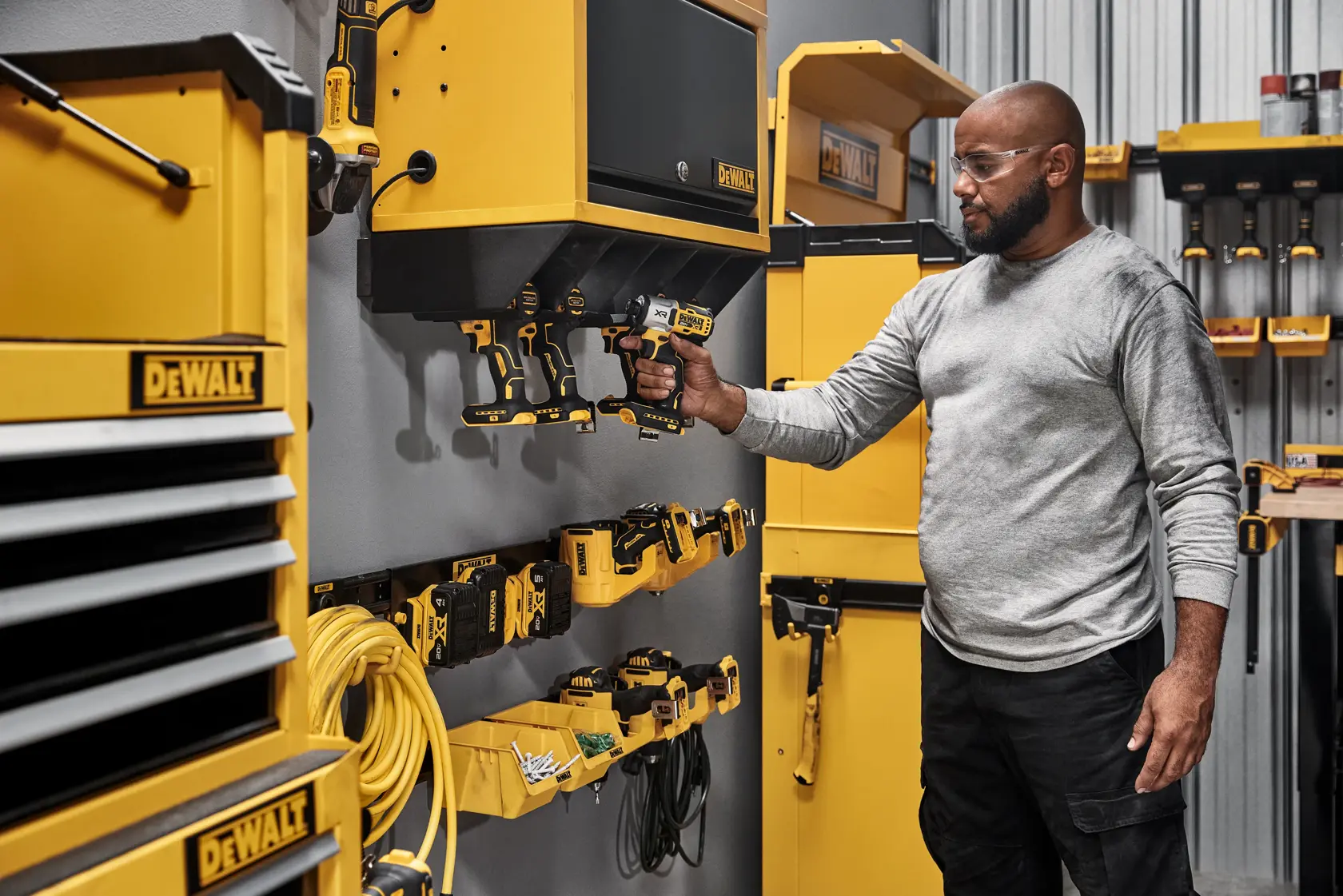 A person in a workshop environment is holding a DEWALT cordless power tool that is stored on a yellow wall-mounted organizer. The workshop has multiple DEWALT branded storage units, tools, and accessories neatly arranged on the walls and shelves.