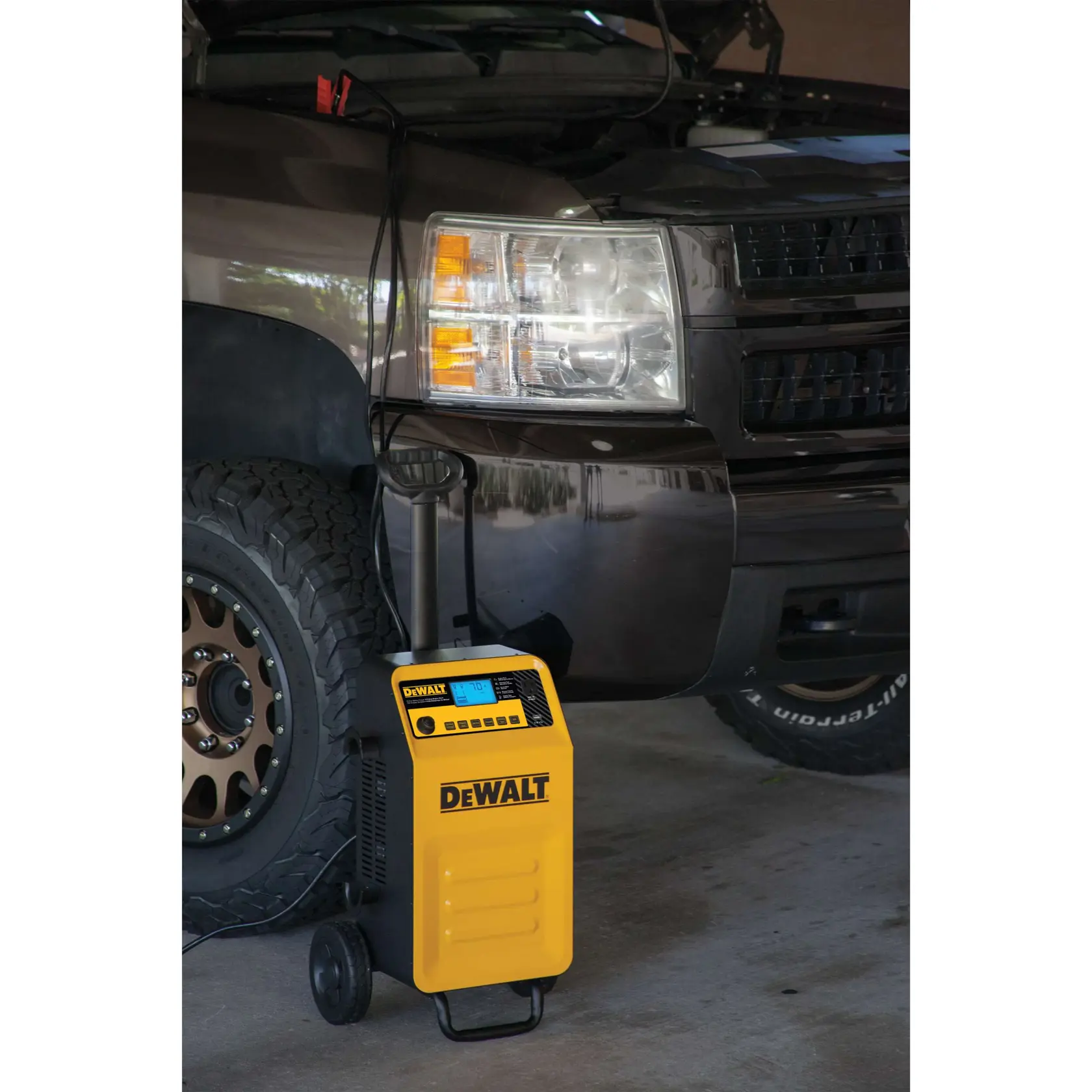 A yellow DEWALT portable battery charger is connected to a vehicle. The charger is positioned on the ground near the front wheel with its cables attached to the car's engine, which has its hood open.