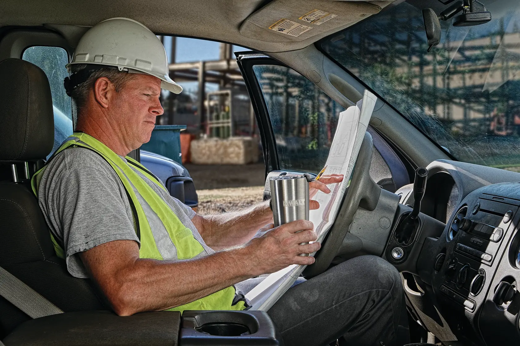A construction worker wearing a safety vest and hard hat sits in the driver's seat of a vehicle, holding blueprints and a DEWALT travel mug.