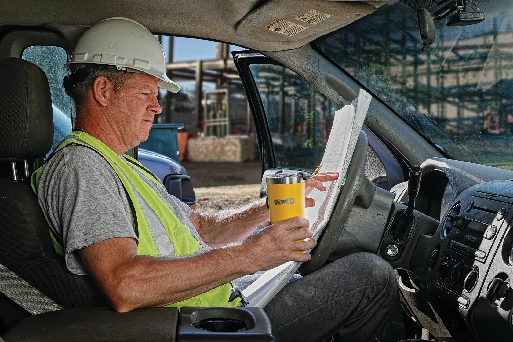 A person wearing a hard hat and safety vest sits in a vehicle, holding a yellow DEWALT tumbler and reviewing large construction plans.
