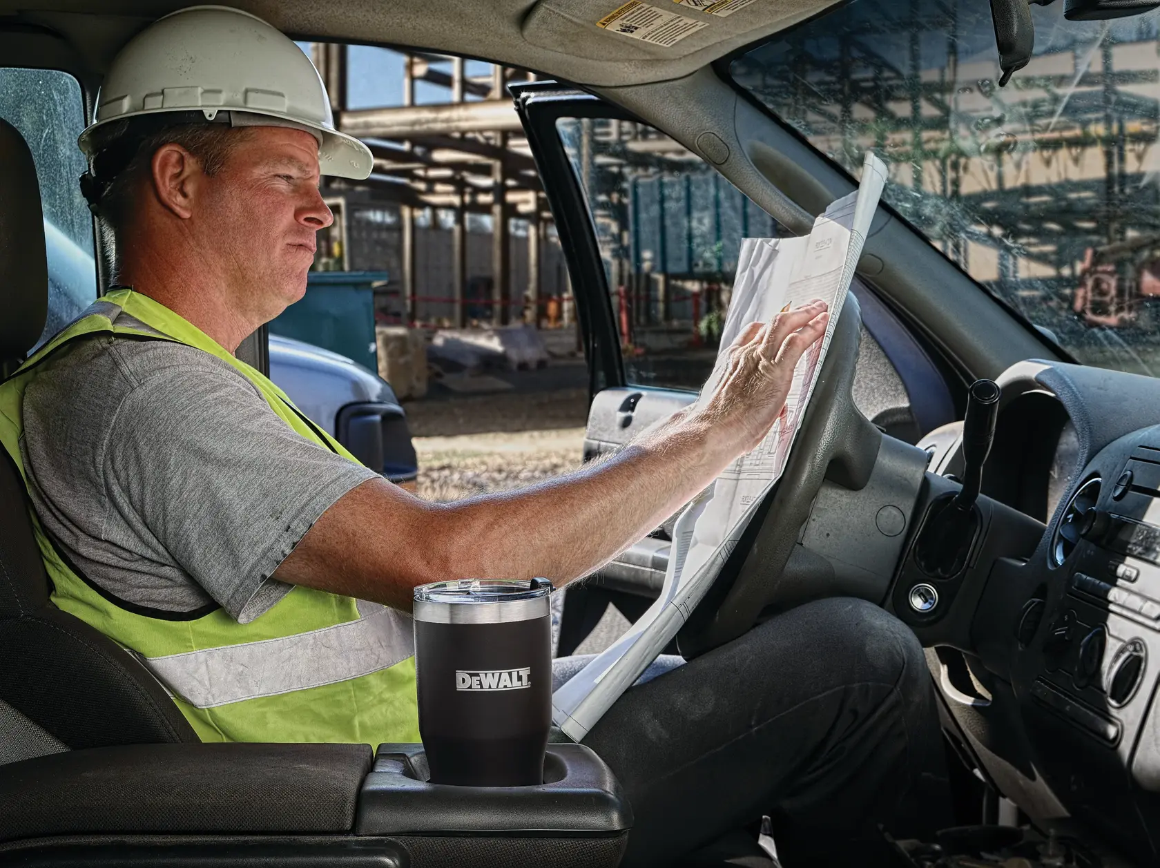 A person wearing a safety vest and hard hat sits in the driver's seat of a vehicle at a construction site, looking at a large paper blueprint. A black DEWALT tumbler is placed in the cup holder.