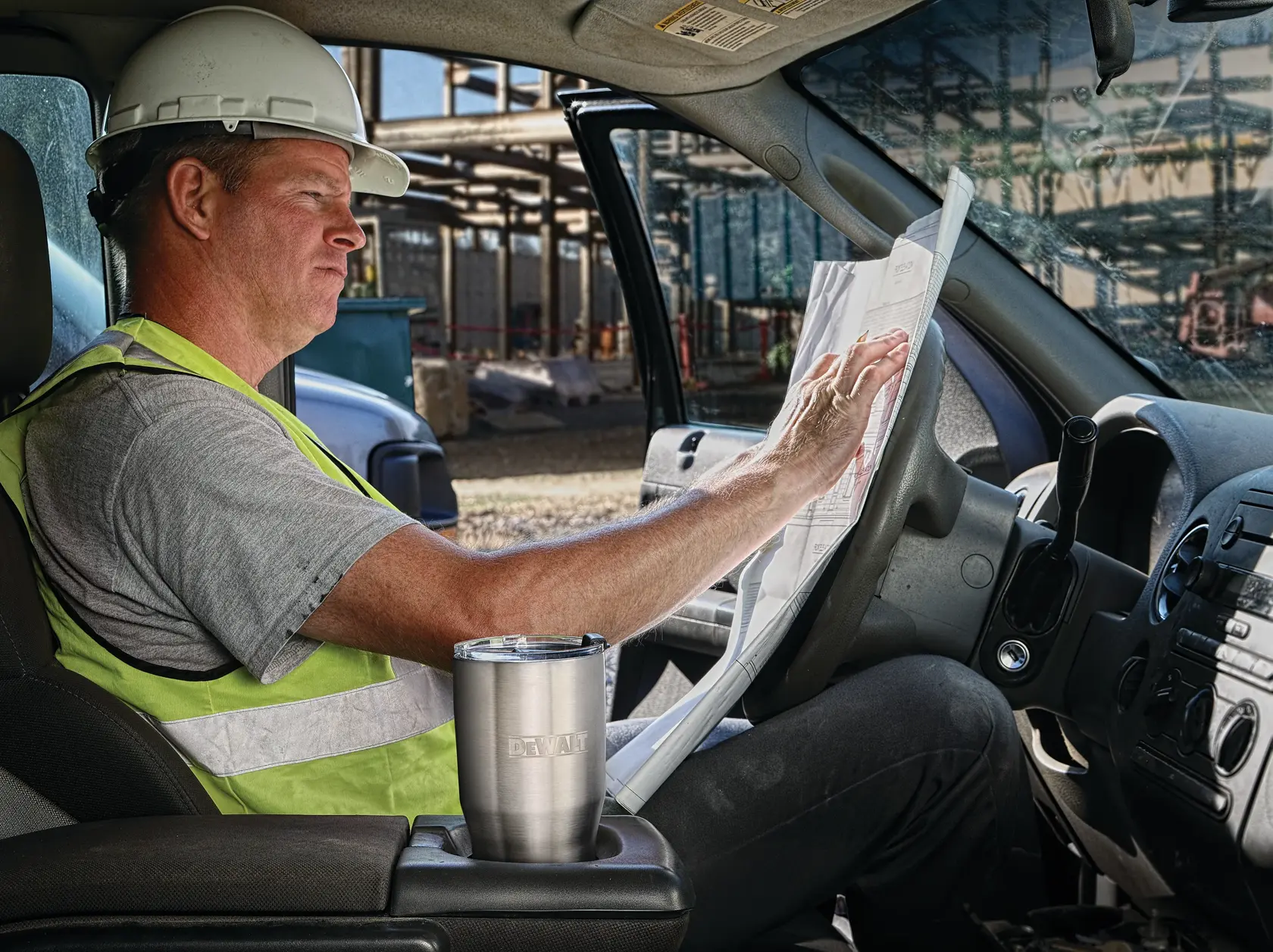 A person wearing a hard hat and safety vest sits in a vehicle and reviews construction plans. A stainless steel DEWALT tumbler is placed in the cup holder. The scene appears to be at a construction site.