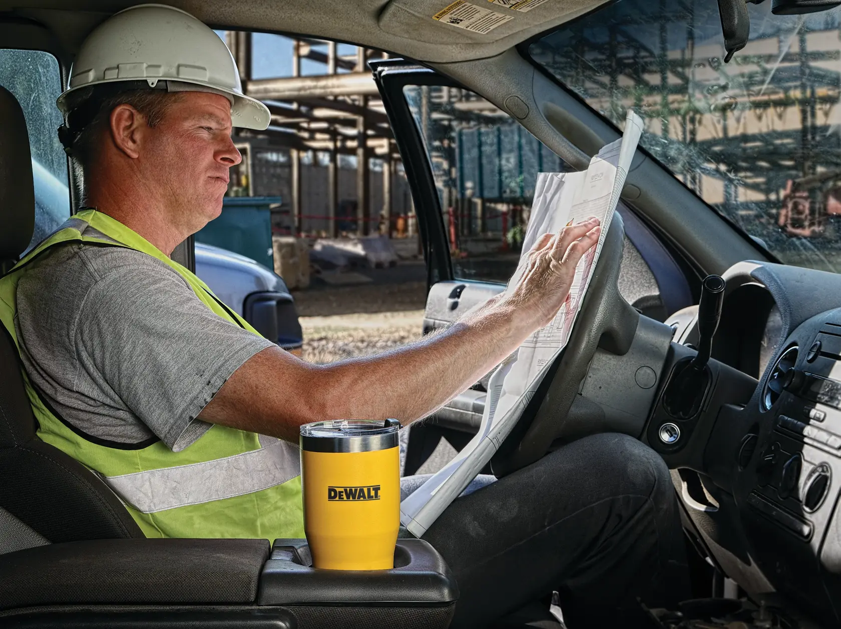 A person wearing a hard hat and safety vest sits in a vehicle, looking at construction plans. A yellow DEWALT travel mug is placed in the cup holder. The background shows a construction site with building structures.