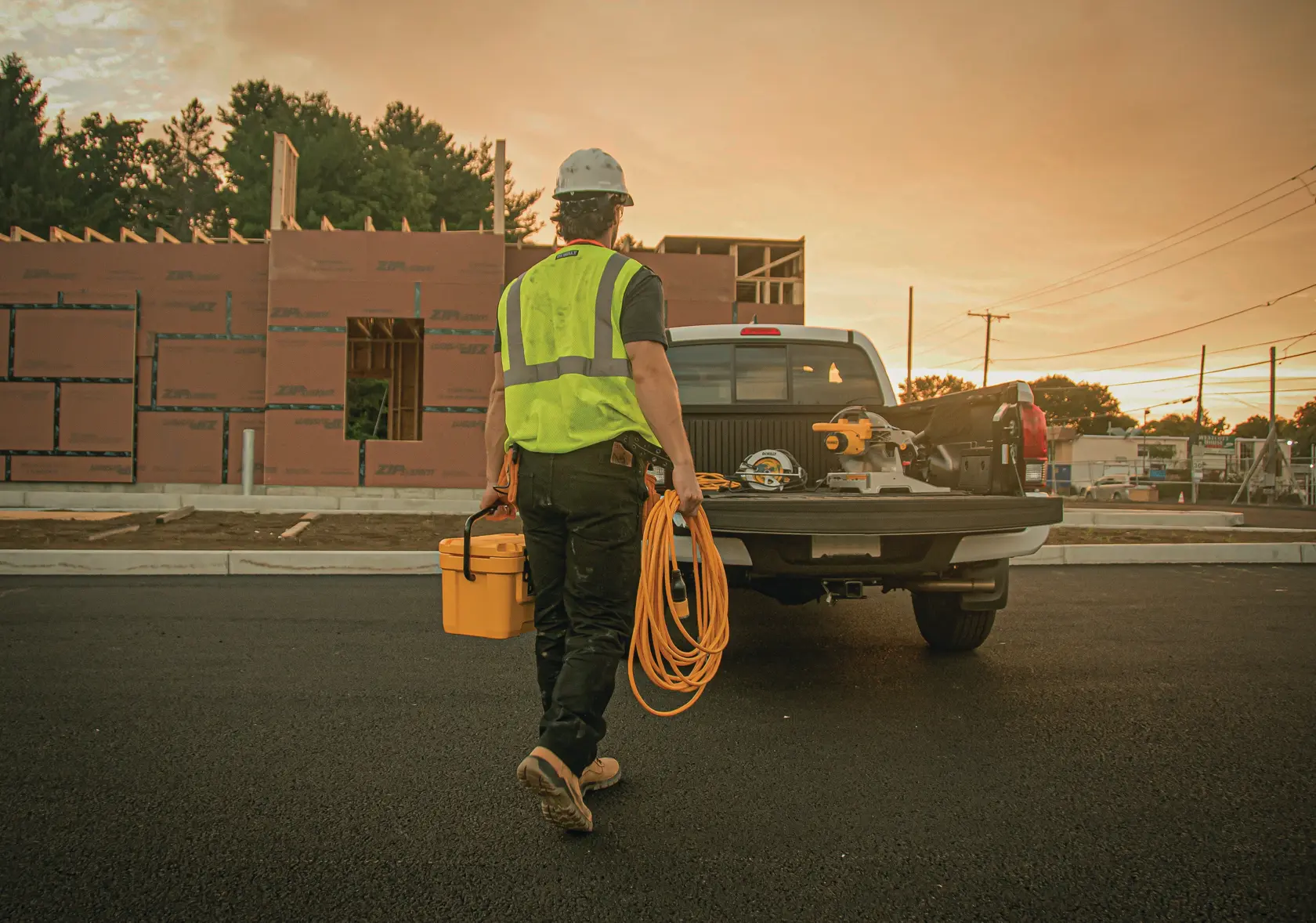 A construction worker wearing a safety vest and helmet walks toward a pickup truck parked at a construction site, carrying a yellow tool box and an orange extension cord. The truck has tools and equipment in the bed, and a partially built structure is visible in the background during sunset.