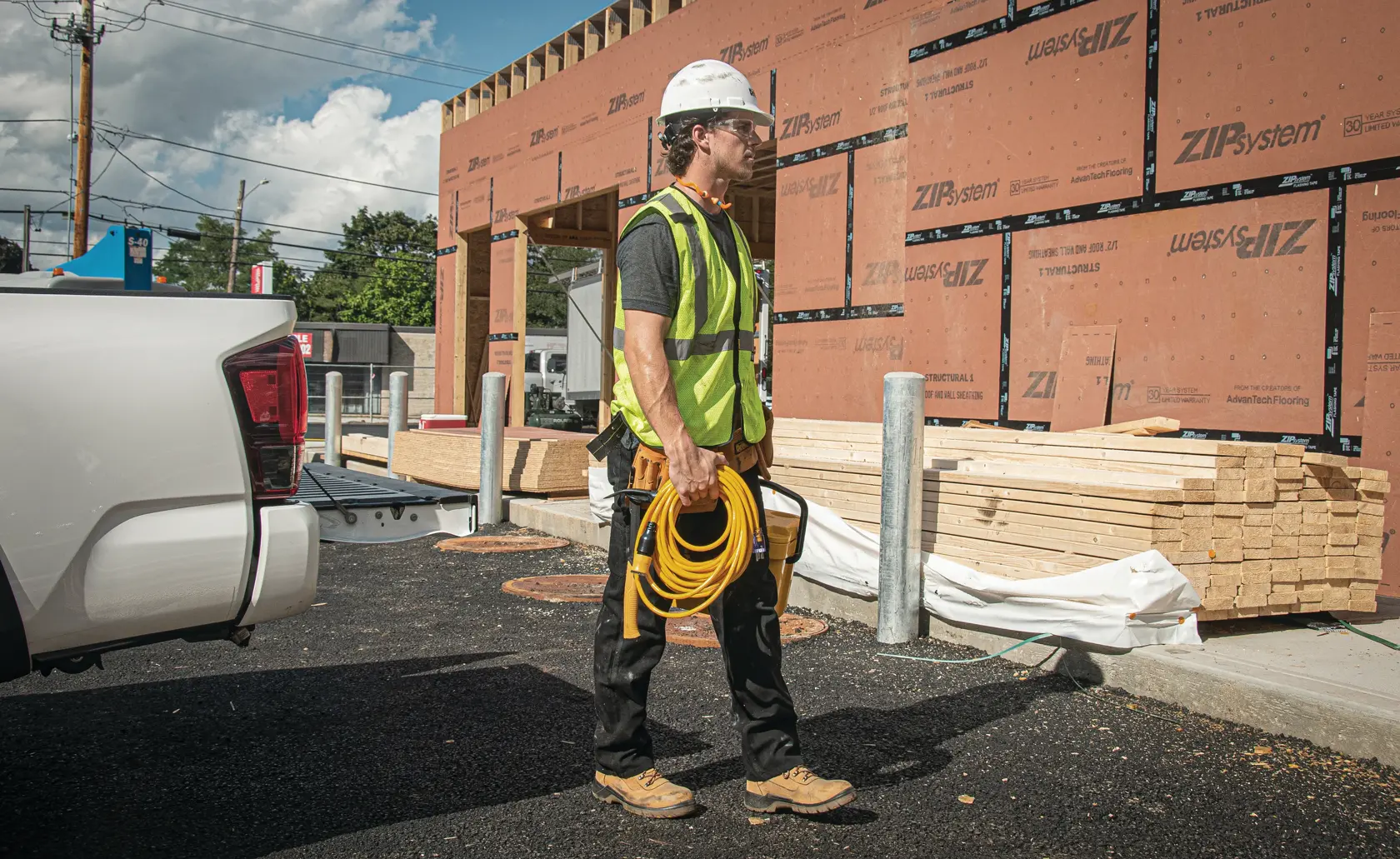 A construction worker wearing a safety vest and hard hat stands outside a building under construction, holding a yellow DEWALT extension cord. Wooden planks and construction materials are visible in the background.