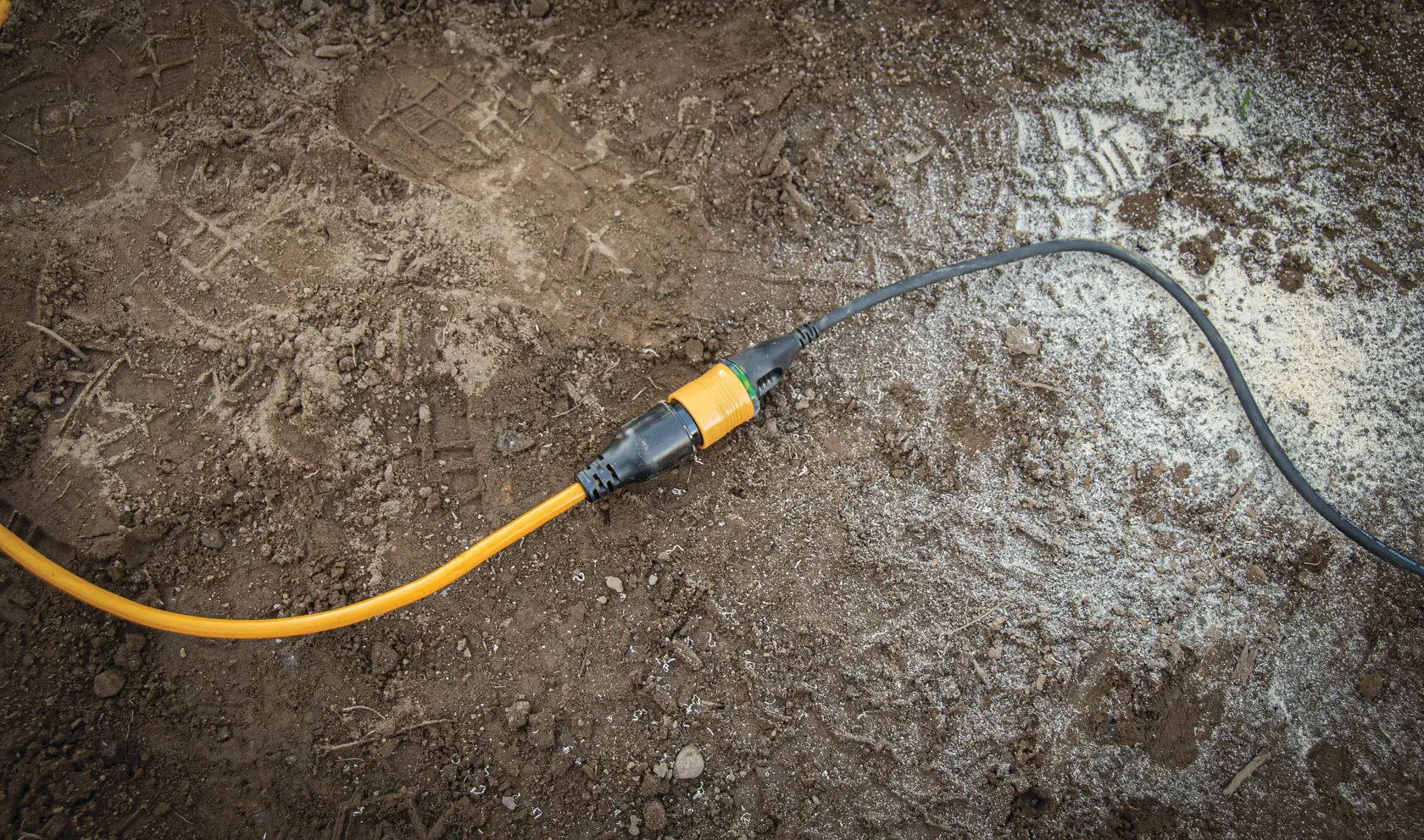 A yellow extension cord connected to a black power cord is lying on a dusty, dirt-covered ground, likely at a construction or work site.