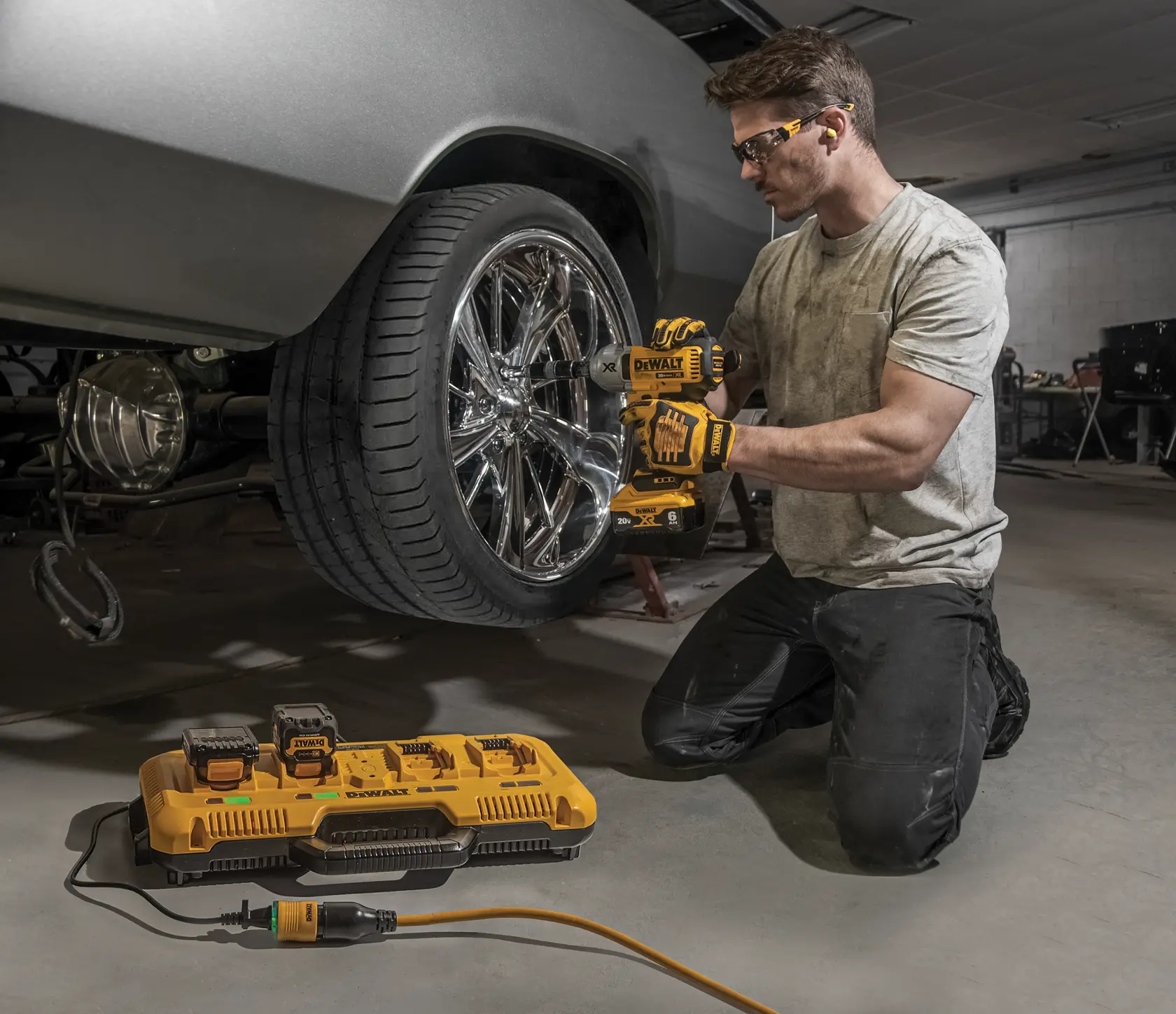 A person kneeling in a garage is using a DEWALT cordless impact wrench to tighten or loosen the lug nuts on a car's shiny chrome wheel. In the foreground, there is a DEWALT battery charging station with two batteries connected, plugged into a power source.