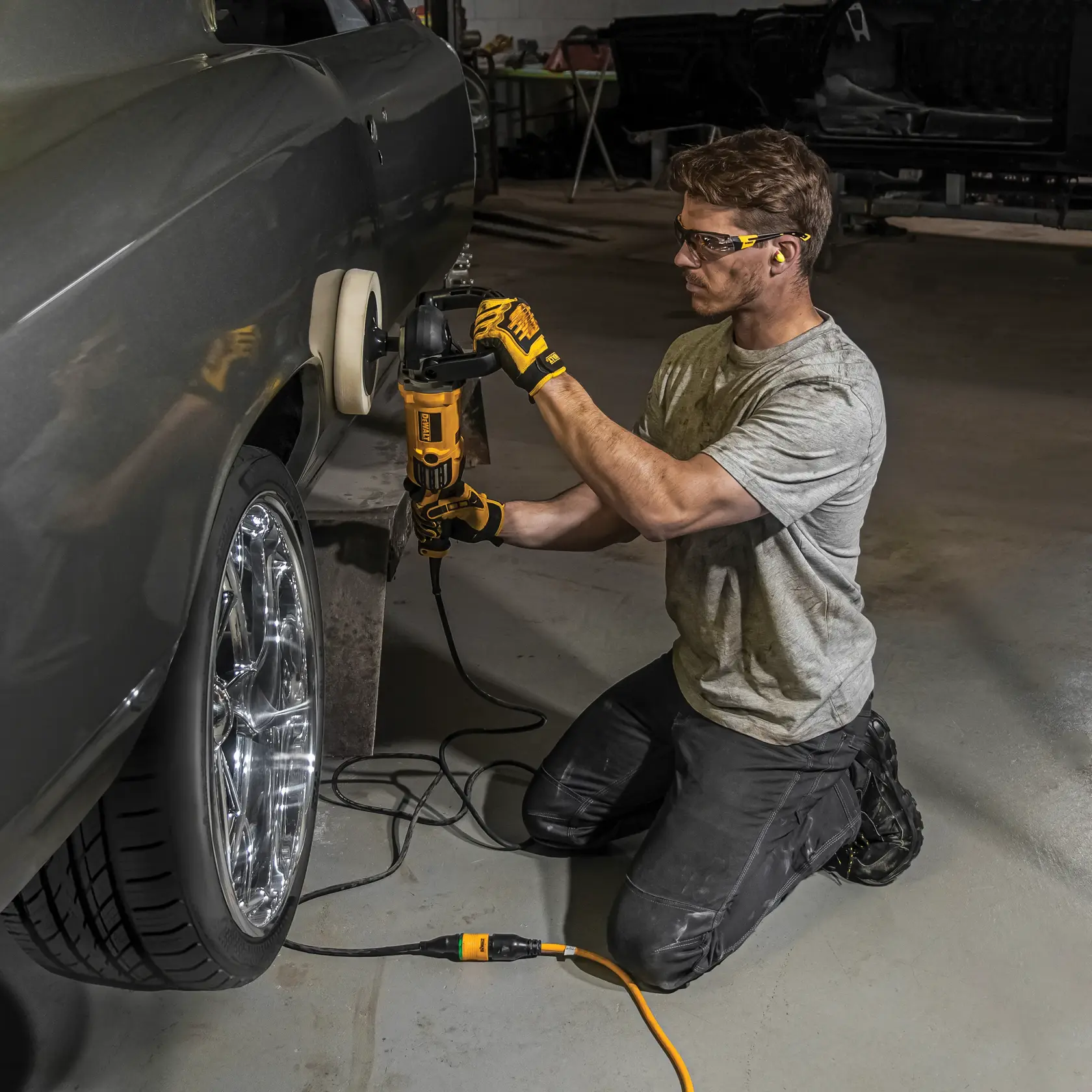 A person kneeling on the floor uses a DEWALT electric polisher to polish the side of a car in a garage. The individual is wearing gloves and ear protection.