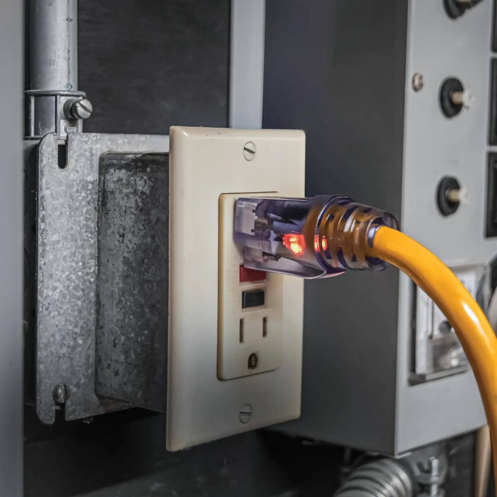 Close-up of a yellow electrical extension cord with a transparent plug connected to a wall outlet. The plug is illuminated with red indicator lights, suggesting active power. The outlet is surrounded by metal and electrical panels.