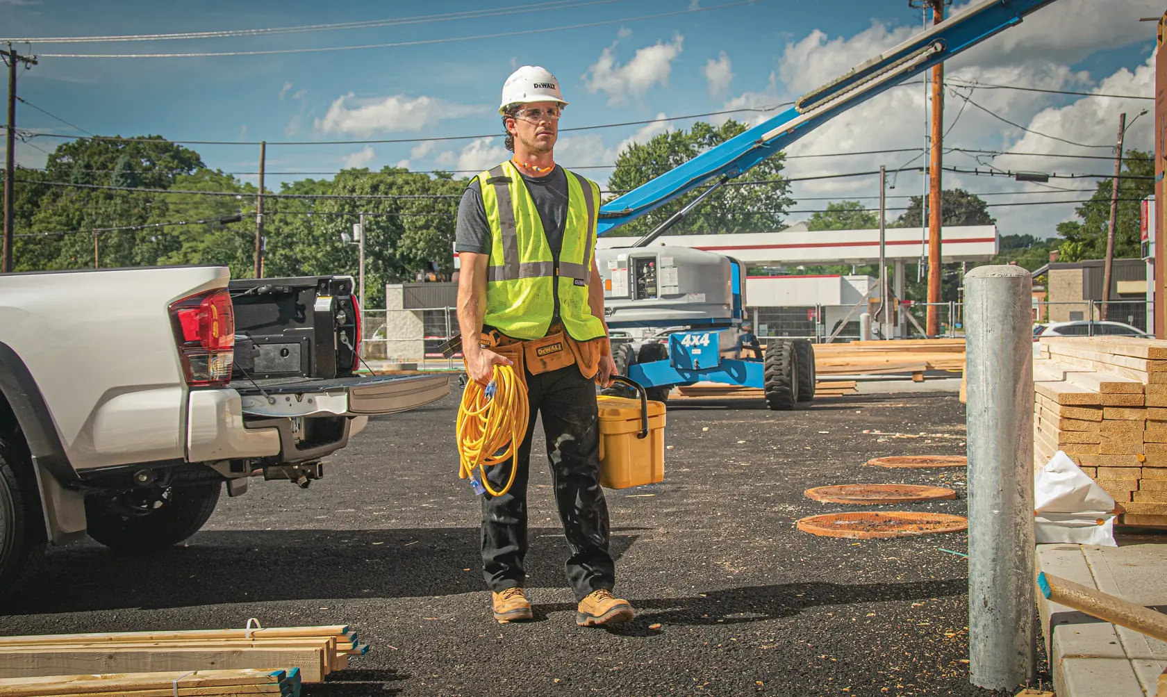 A construction worker wearing a high-visibility vest and hard hat stands outdoors near a white pickup truck. The worker is holding a yellow extension cord and a yellow toolbox. Construction materials and equipment are visible in the background.