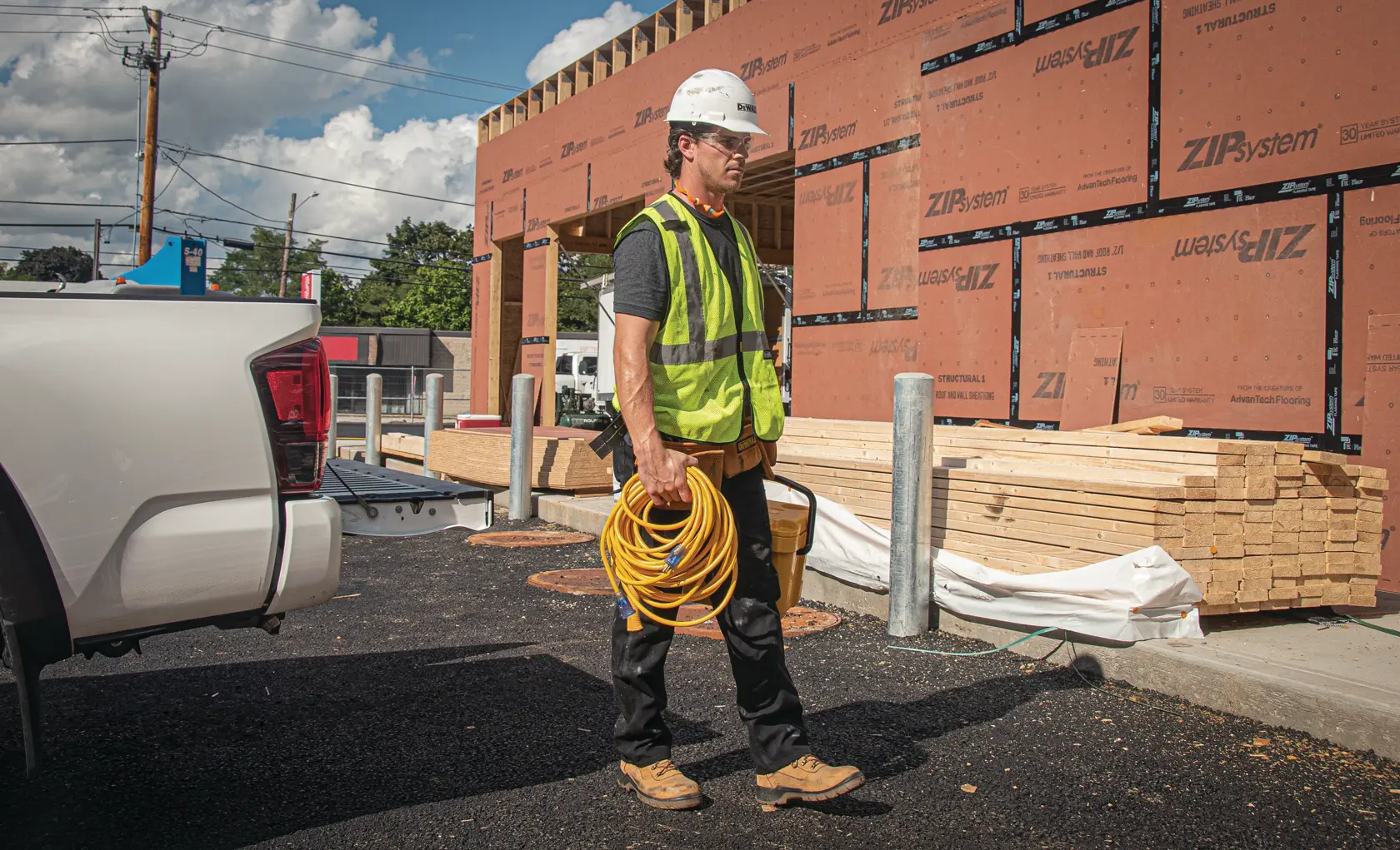 A construction worker wearing a yellow safety vest and white helmet stands near a construction site, holding a yellow extension cord. Lumber and building materials are stacked beside a partially built structure with ZIP System sheathing. A white pickup truck is visible on the left.