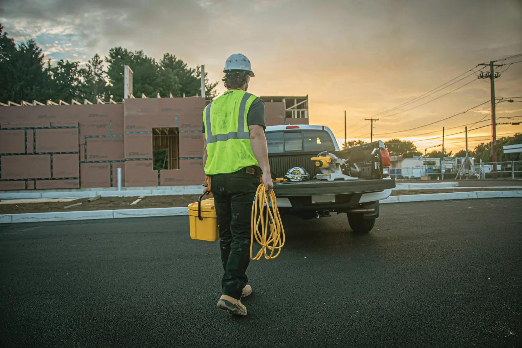 A construction worker wearing a yellow safety vest and helmet is walking towards a pickup truck with building tools and equipment in the truck bed. The worker is holding a yellow toolbox and an extension cord, with a construction site and unfinished building in the background during sunset.