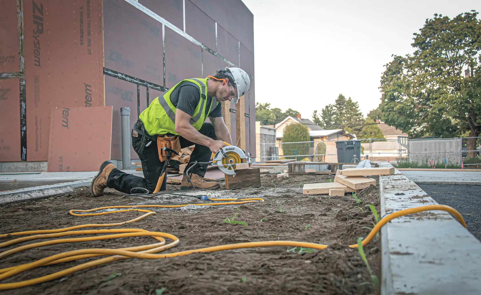 A construction worker wearing a safety vest and helmet uses a DEWALT circular saw to cut wood at an outdoor job site. Yellow extension cords are visible on the ground, and the work area is near a partially built structure.
