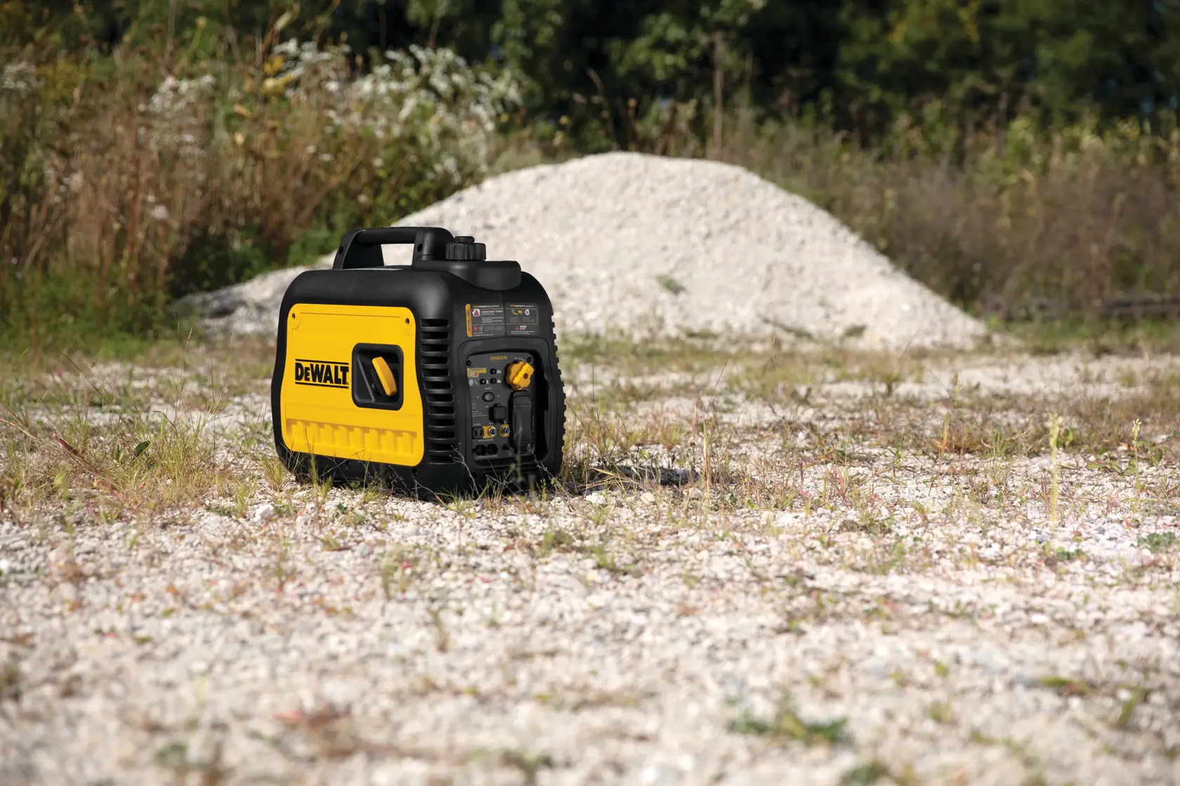 A portable DEWALT inverter generator, model DXGNI2200, is placed outdoors on a gravel surface with grass and a pile of stones in the background.