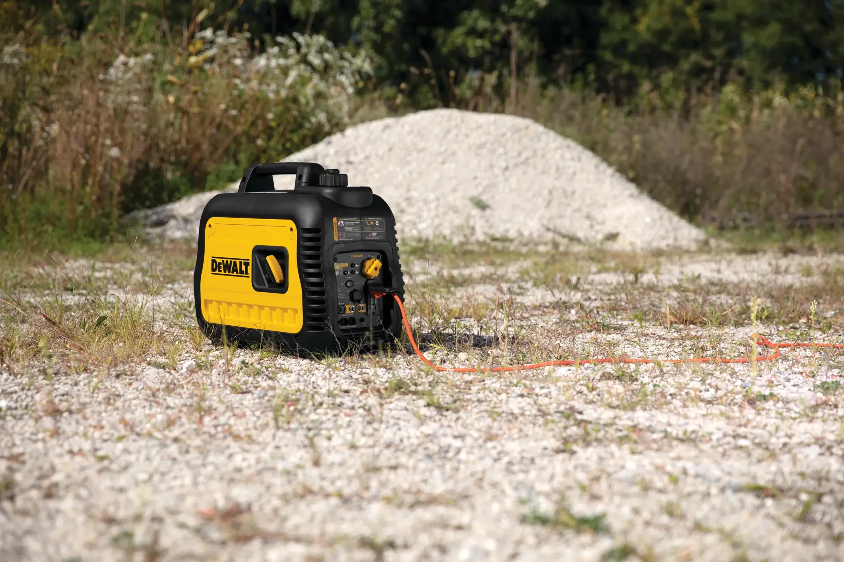 A DEWALT portable generator is placed outdoors on a gravel surface with some grass, near a mound of white rocks, and an orange extension cord is plugged into it.