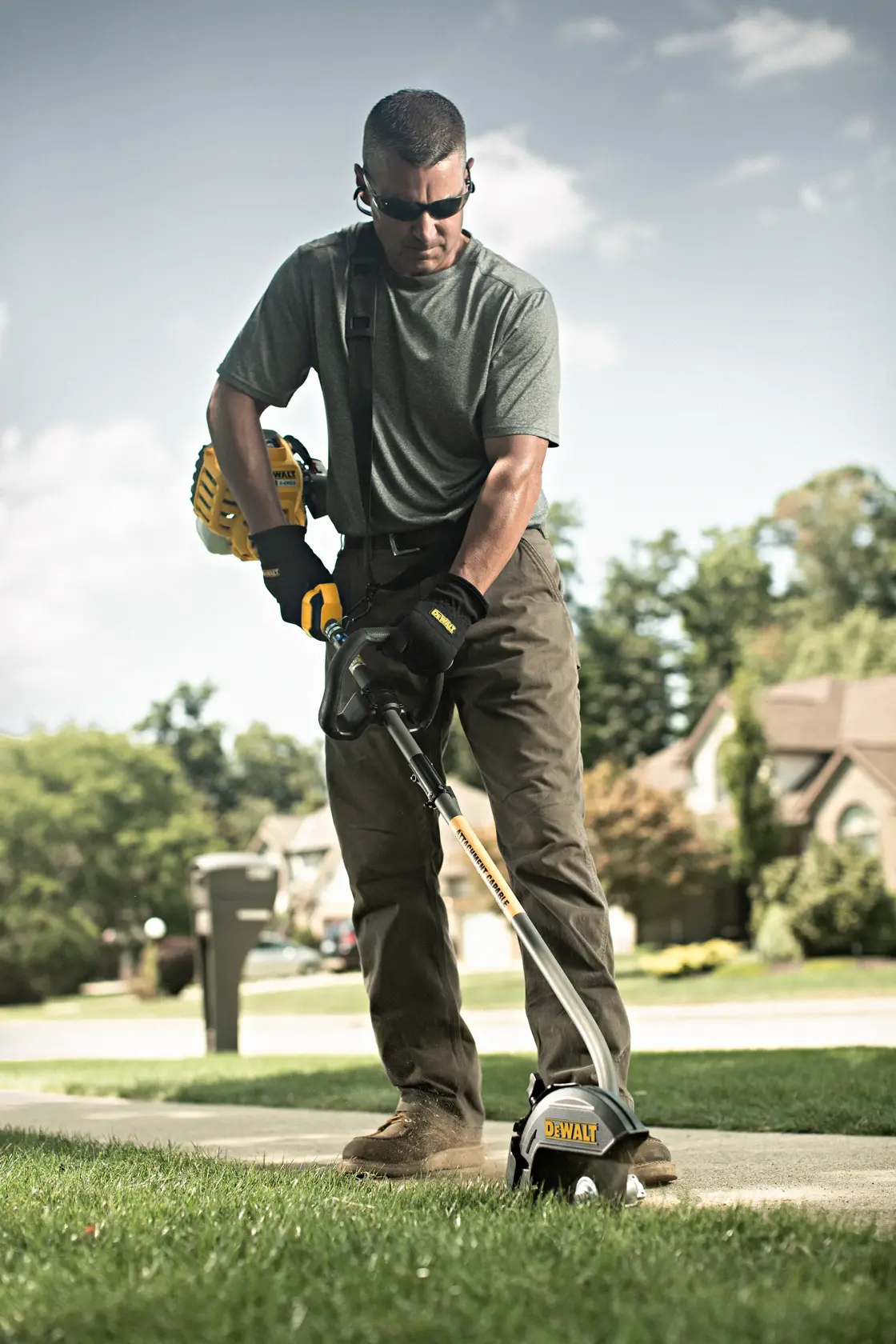 A person is using a DEWALT outdoor power tool to edge the grass along a sidewalk in a residential area. The individual is wearing gloves, work pants, and boots, with houses and greenery visible in the background.