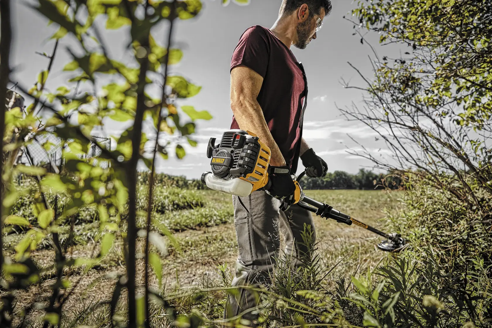 A person outdoors operating a DEWALT gas-powered string trimmer, cutting grass and weeds near bushes and trees. The person is wearing gloves and a maroon shirt, with their face blurred.