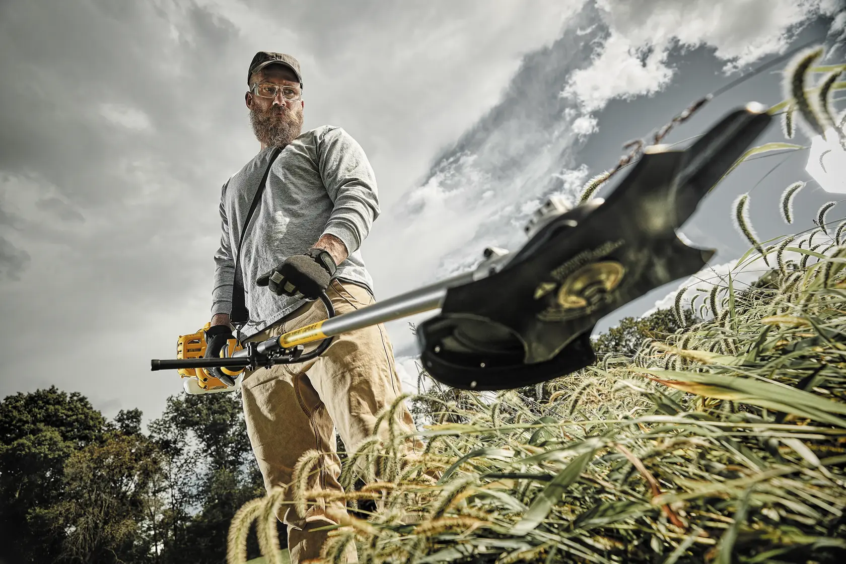 A person standing outdoors using a DEWALT string trimmer, cutting tall grass. The person is wearing protective gloves and casual clothing. The sky is cloudy with trees in the background. The person's face is not visible.