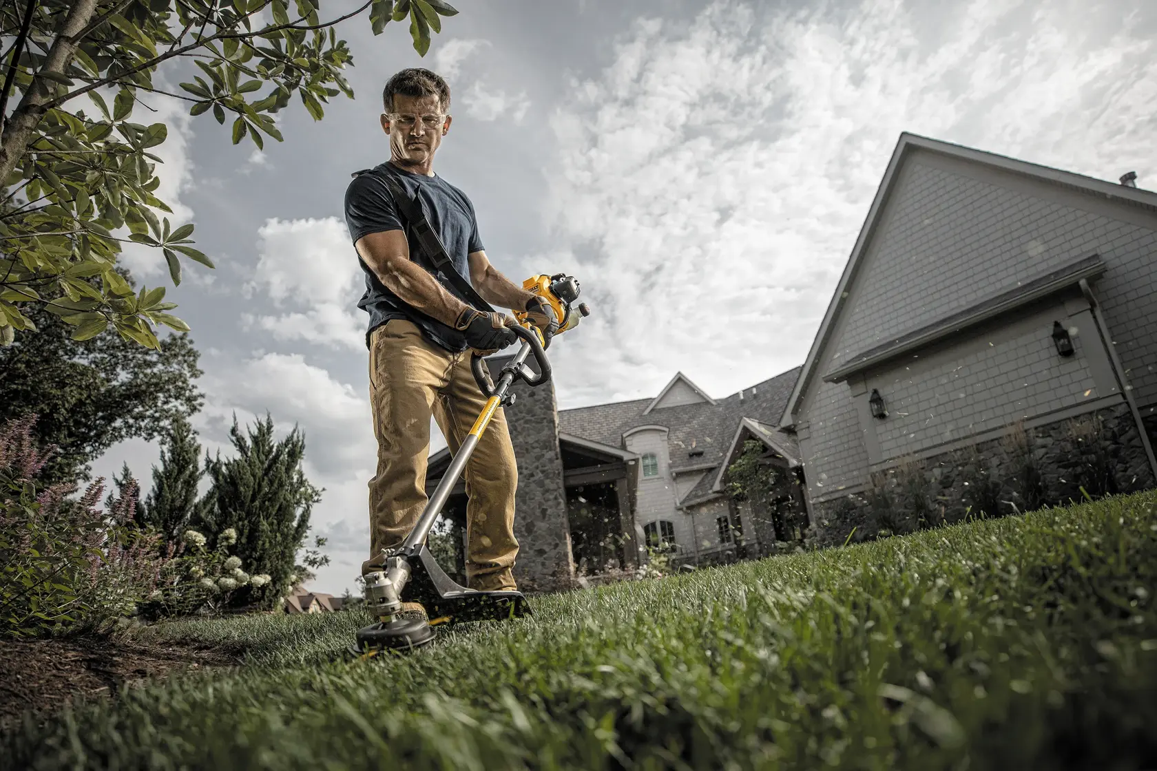 A person using a DEWALT string trimmer to cut grass in a residential yard. The individual is wearing gloves and standing near a house on a sunny day.