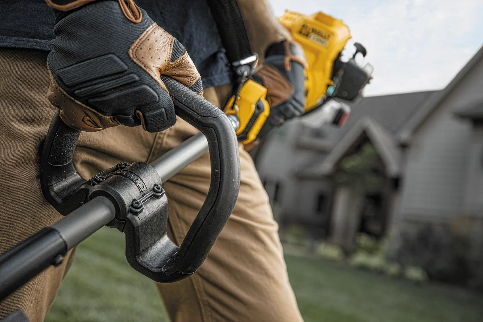 Close-up view of a person wearing gloves and holding a DEWALT outdoor power tool, likely a string trimmer, in a residential yard.