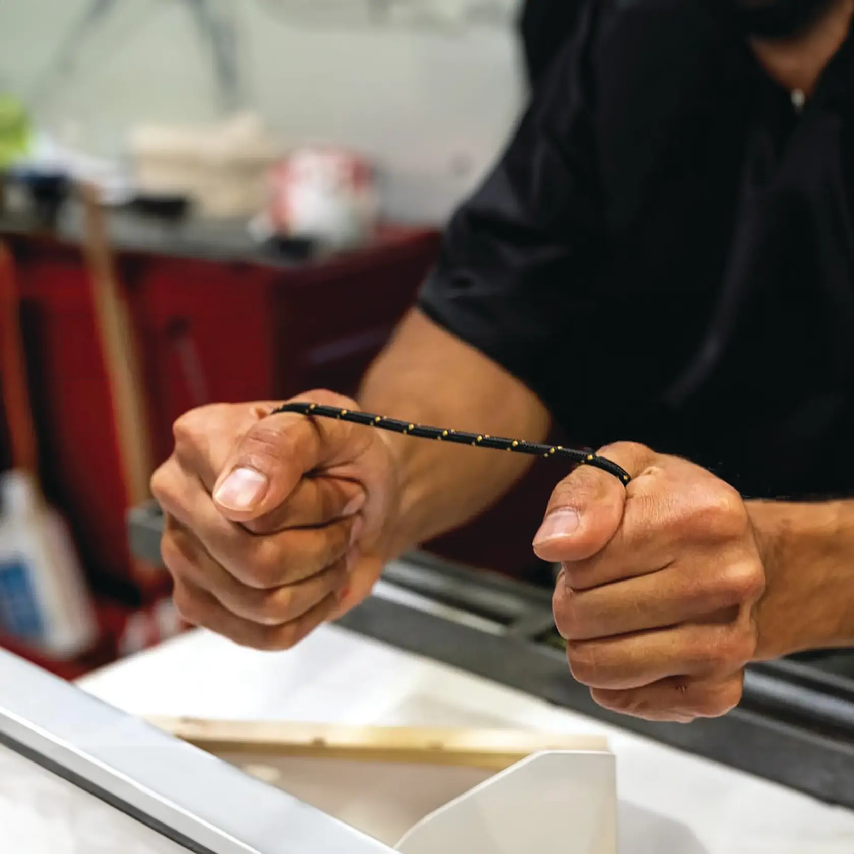 A person holding and stretching a black cord with yellow markings in both hands, possibly in a workshop or workspace.