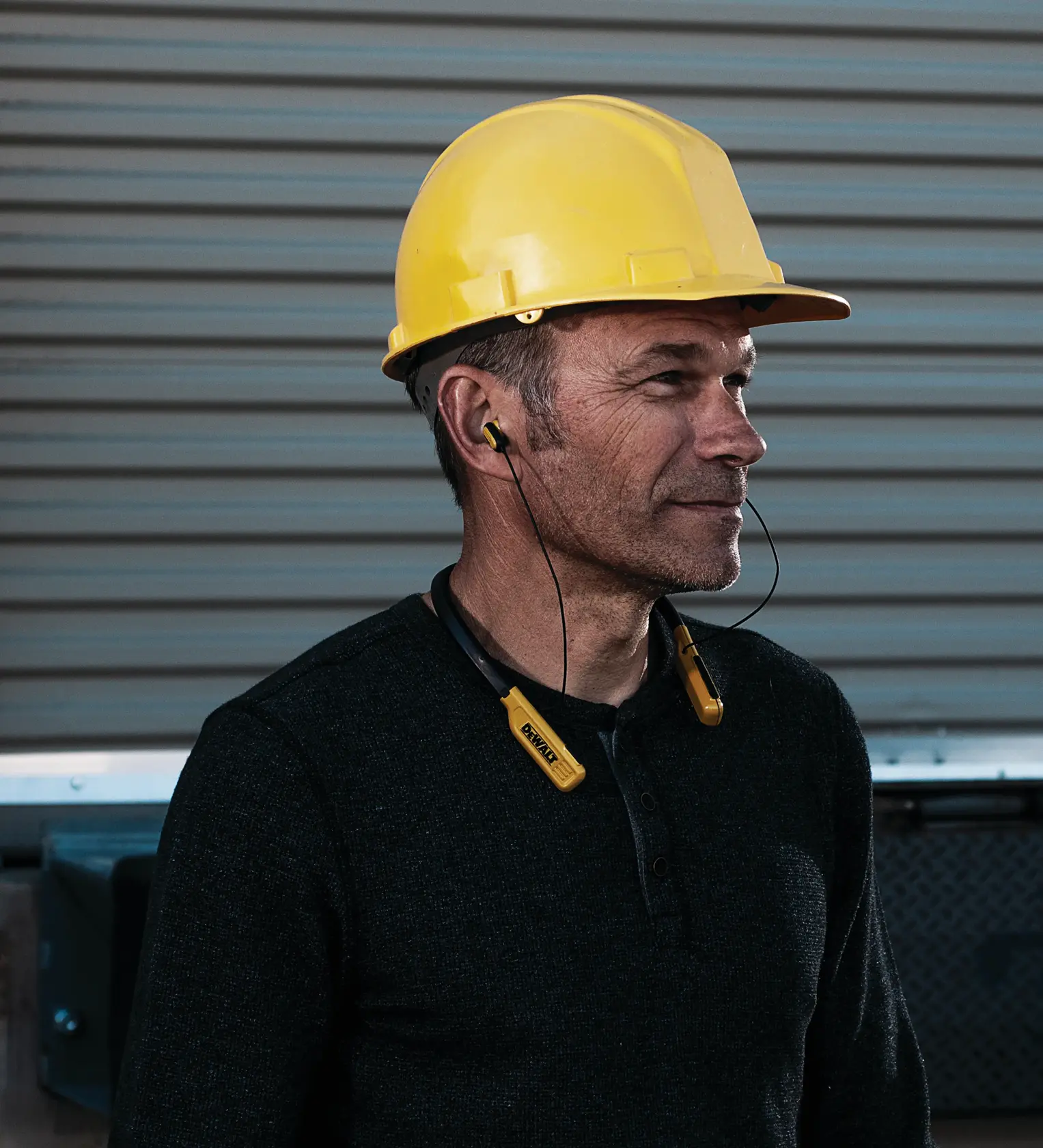 Person wearing a yellow hard hat and black shirt, using DEWALT neckband-style wireless earphones with yellow earbuds, standing in front of a metal garage door.