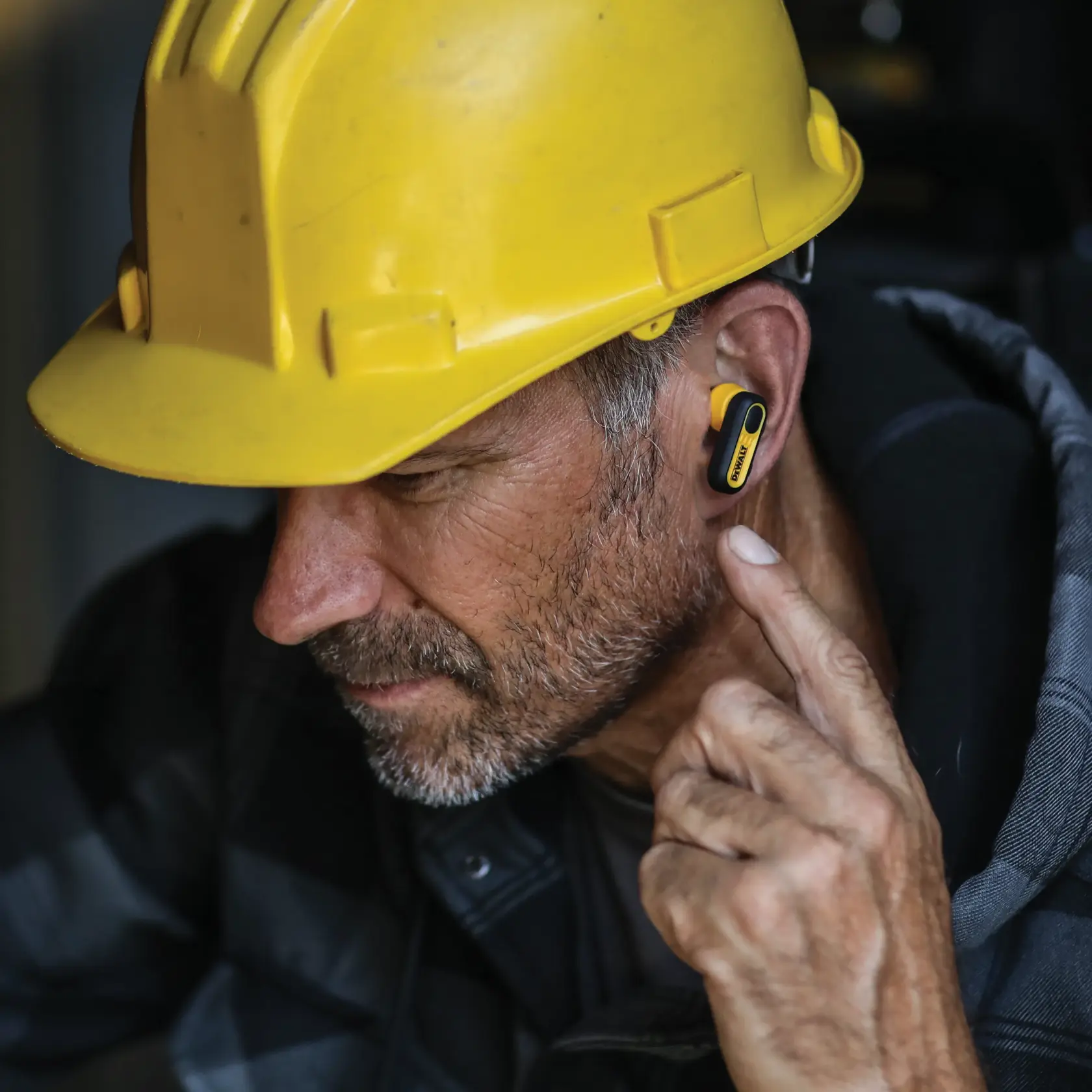 A person wearing a yellow hard hat and a yellow DEWALT earbud, with their finger touching the earbud. The image focuses on the ear protection device, which is suitable for use on worksites.