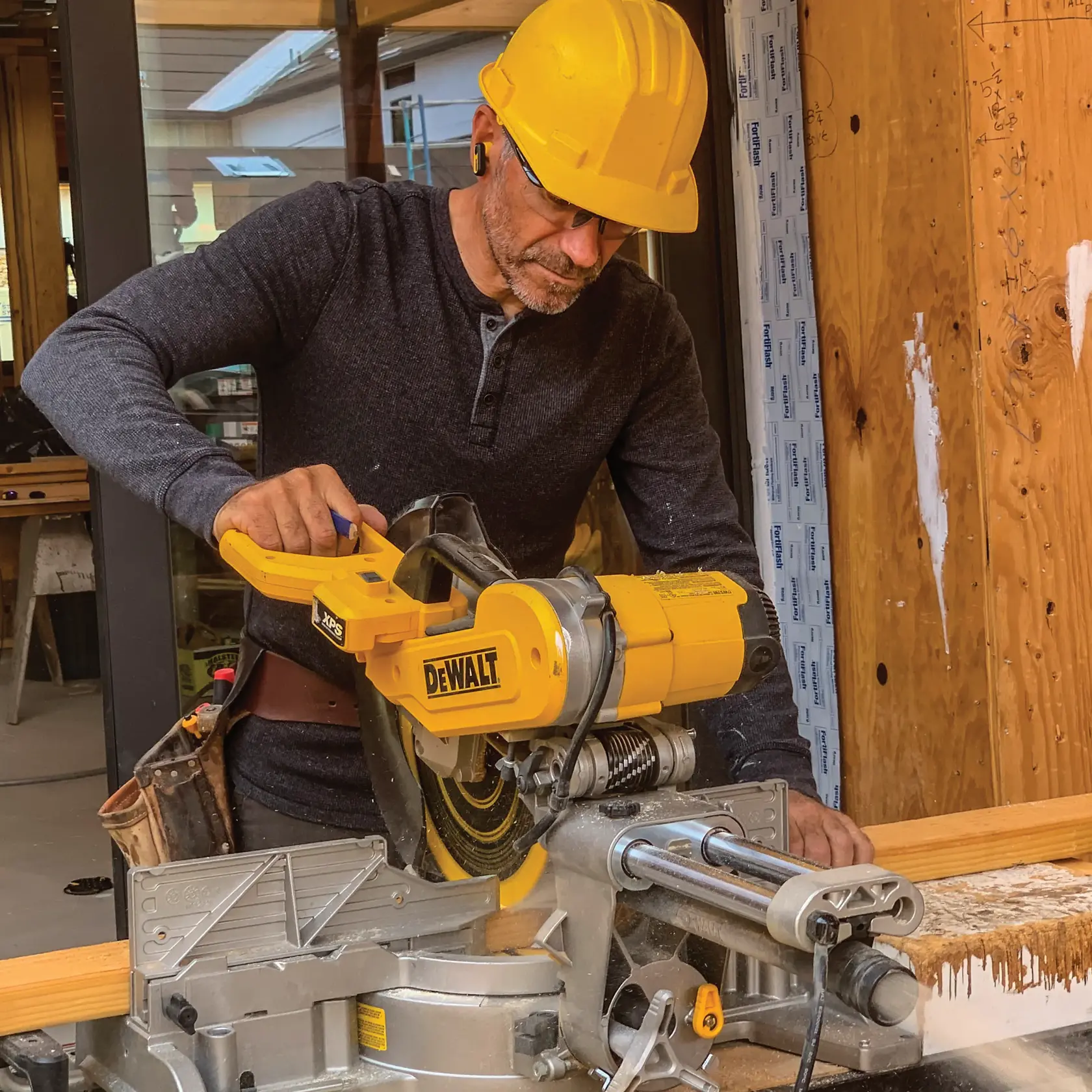 A person wearing a yellow hard hat is using a DEWALT compound miter saw to cut a piece of wood on a construction site. The DEWALT saw is yellow and silver, and the person is dressed in a dark long-sleeve shirt and tool belt.