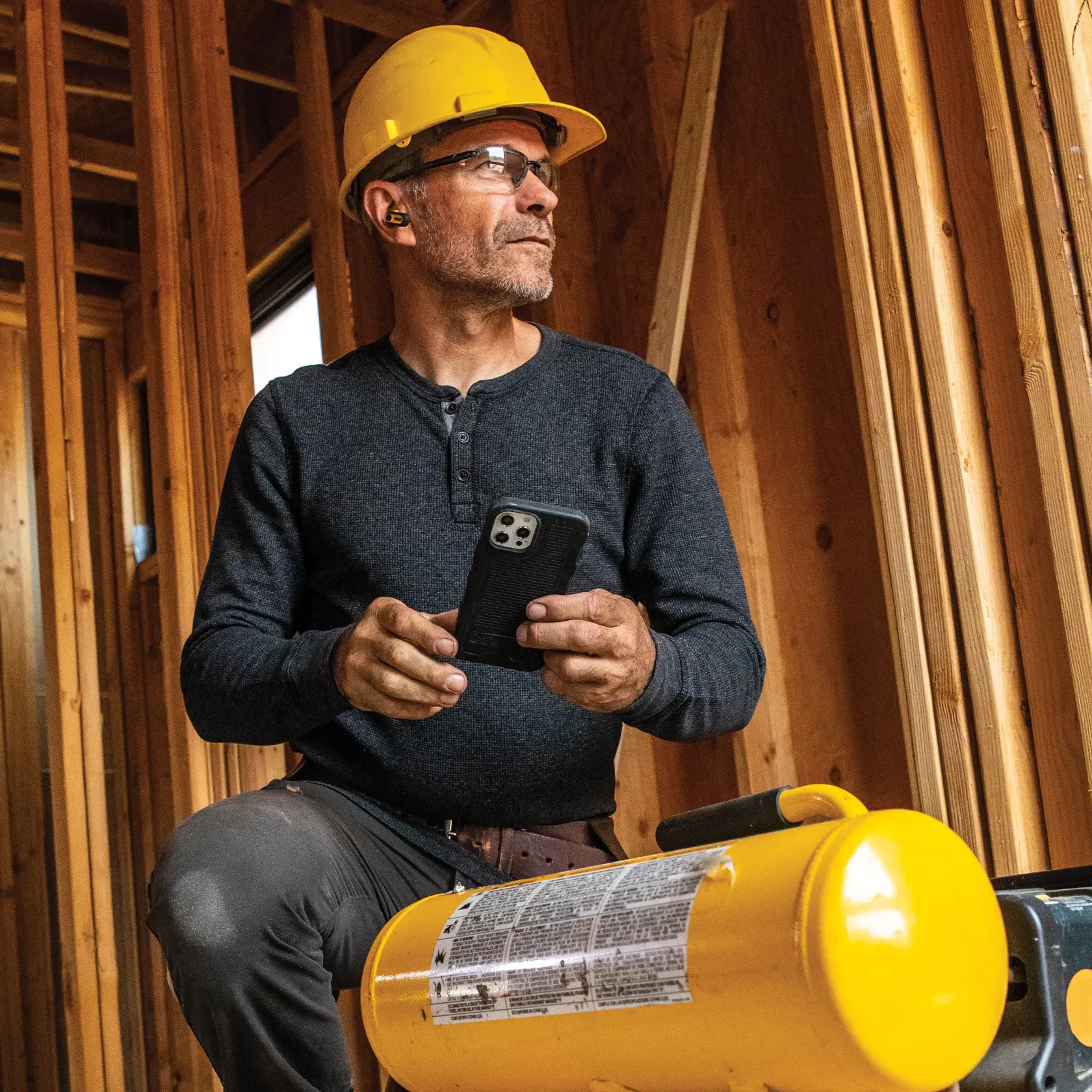 Person wearing a yellow hard hat and ear protection, seated in an unfinished wooden building. The individual is using a smartphone, with a DEWALT-branded yellow device visible in the foreground.