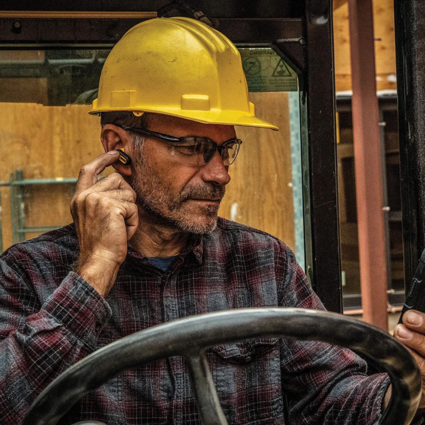 A person wearing a yellow hard hat and a plaid shirt sits in a construction vehicle cab and adjusts a DEWALT wireless earpiece. The person is holding a device in their other hand, with their face obscured for privacy.