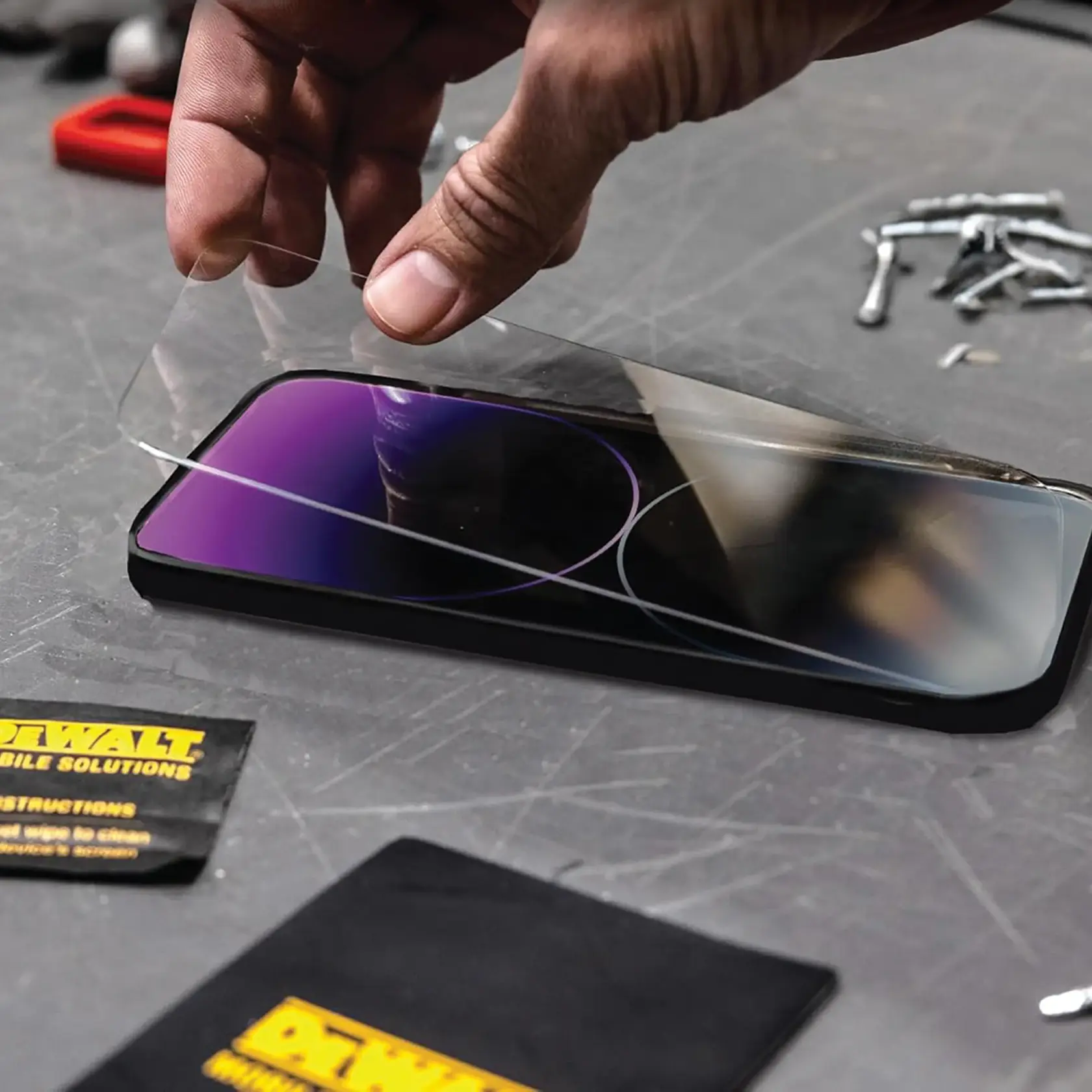 A hand is applying a clear screen protector to a smartphone on a workbench. The scene includes DEWALT branded cleaning packets and other small tools scattered on the surface.