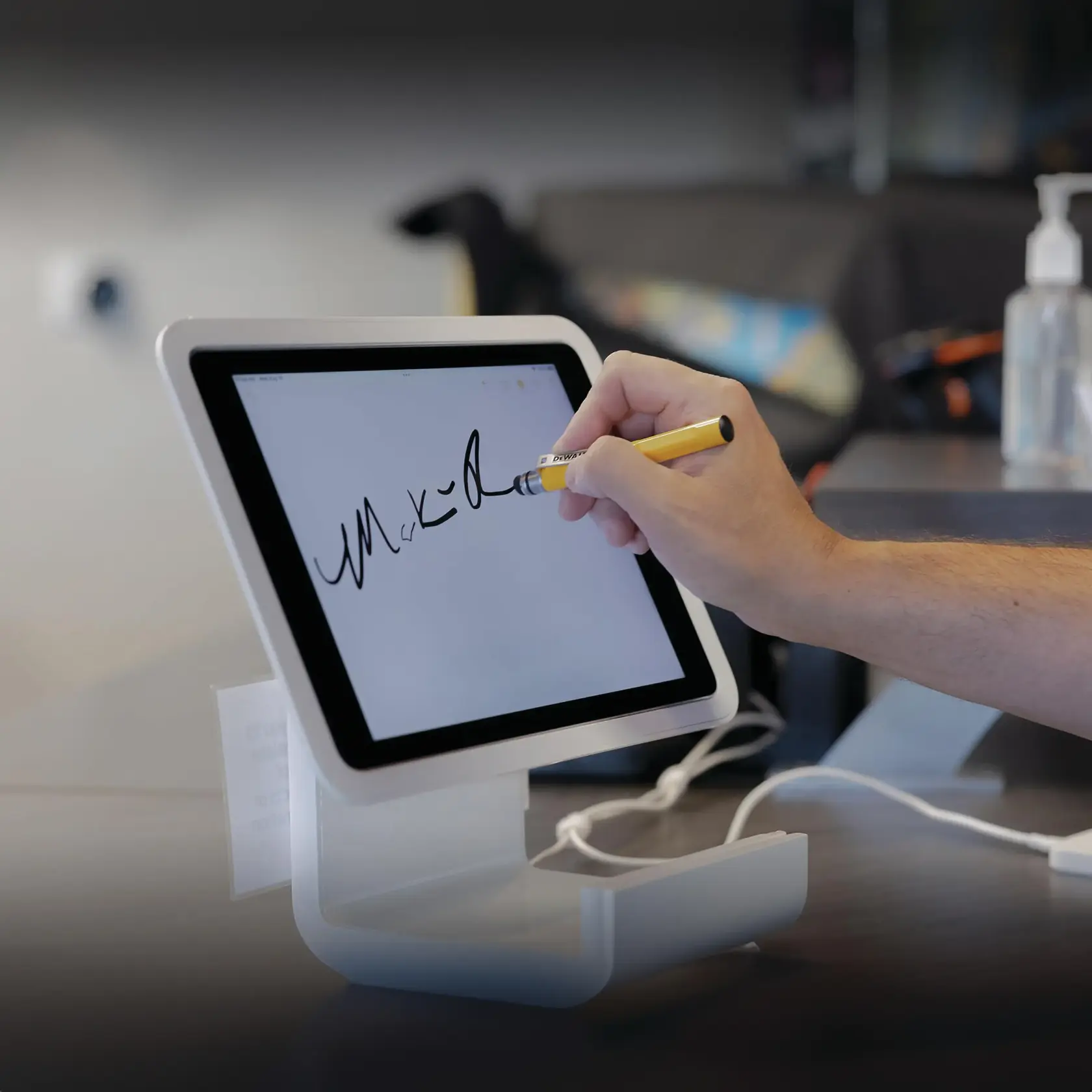 A person using a stylus to sign their name on a tablet that is placed on a stand. The tablet screen displays a handwritten signature, and the environment appears to be an indoor setting, possibly a reception or checkout area.