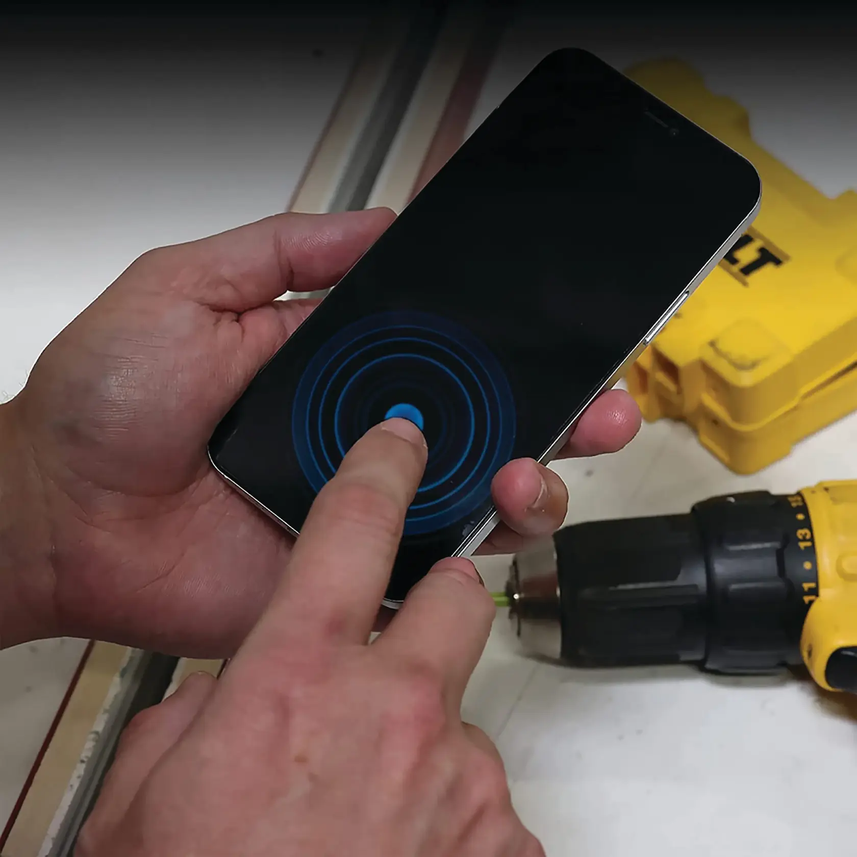 A person is using a smartphone with a circular touch interface displayed on the screen. A yellow DEWALT drill and tool case are visible in the background on a white surface.