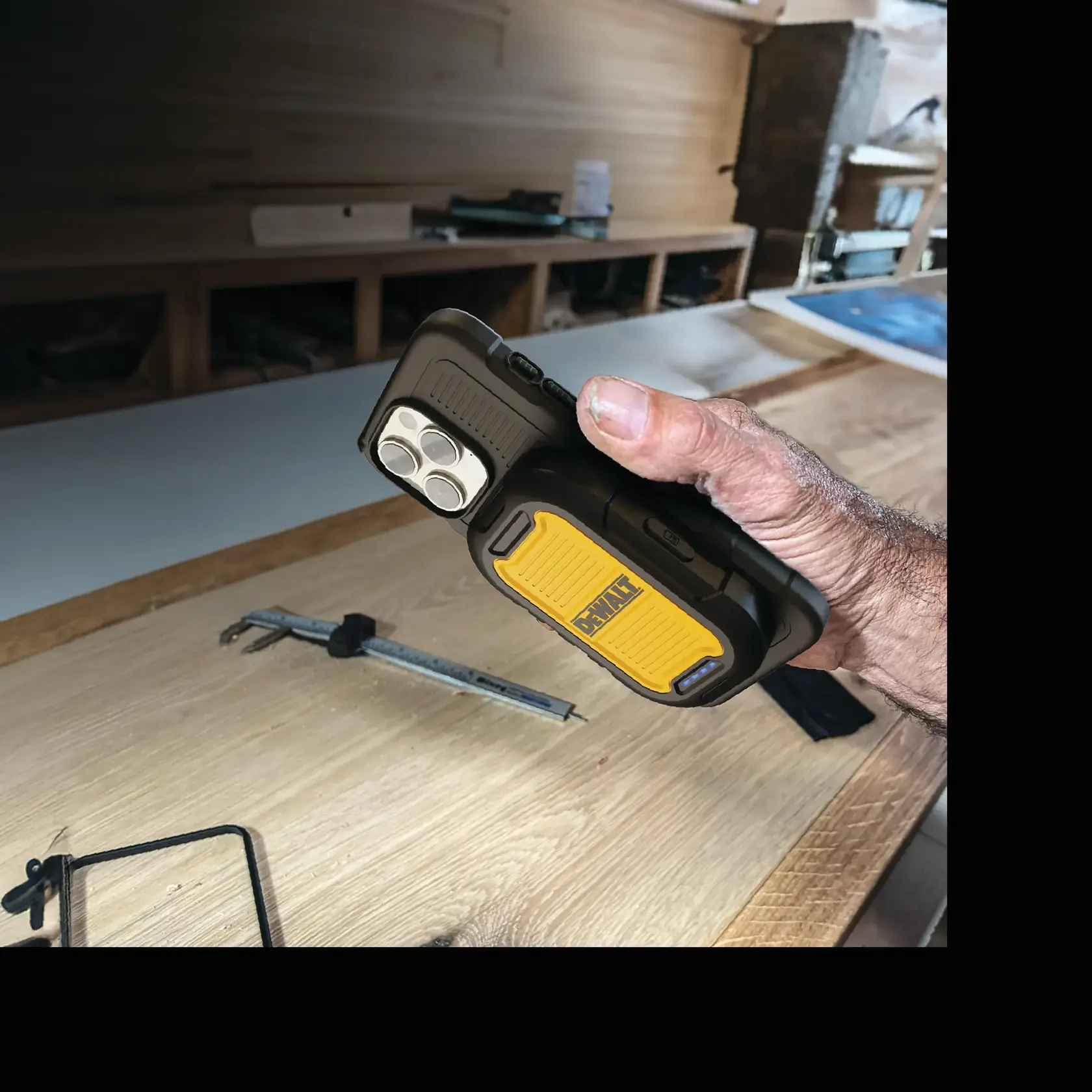 A hand holds a DEWALT-branded rugged smartphone case with a built-in yellow and black attachment, likely designed for worksite environments. The case is being used in a woodworking shop, with tools and wood visible in the background.