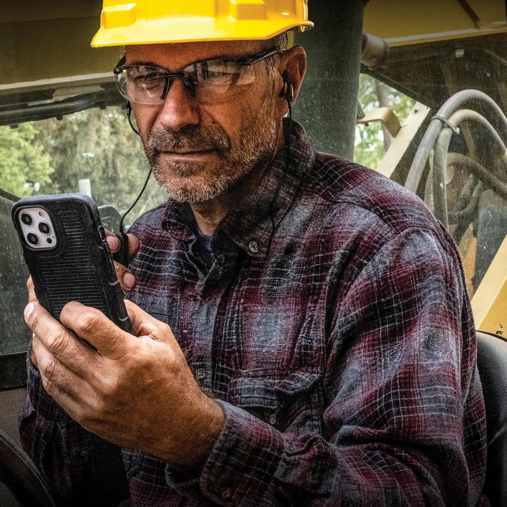 A person wearing a yellow hard hat and a plaid shirt is holding a smartphone in a rugged black case. The individual is seated inside construction machinery and is using wired earphones. Their face is obscured.