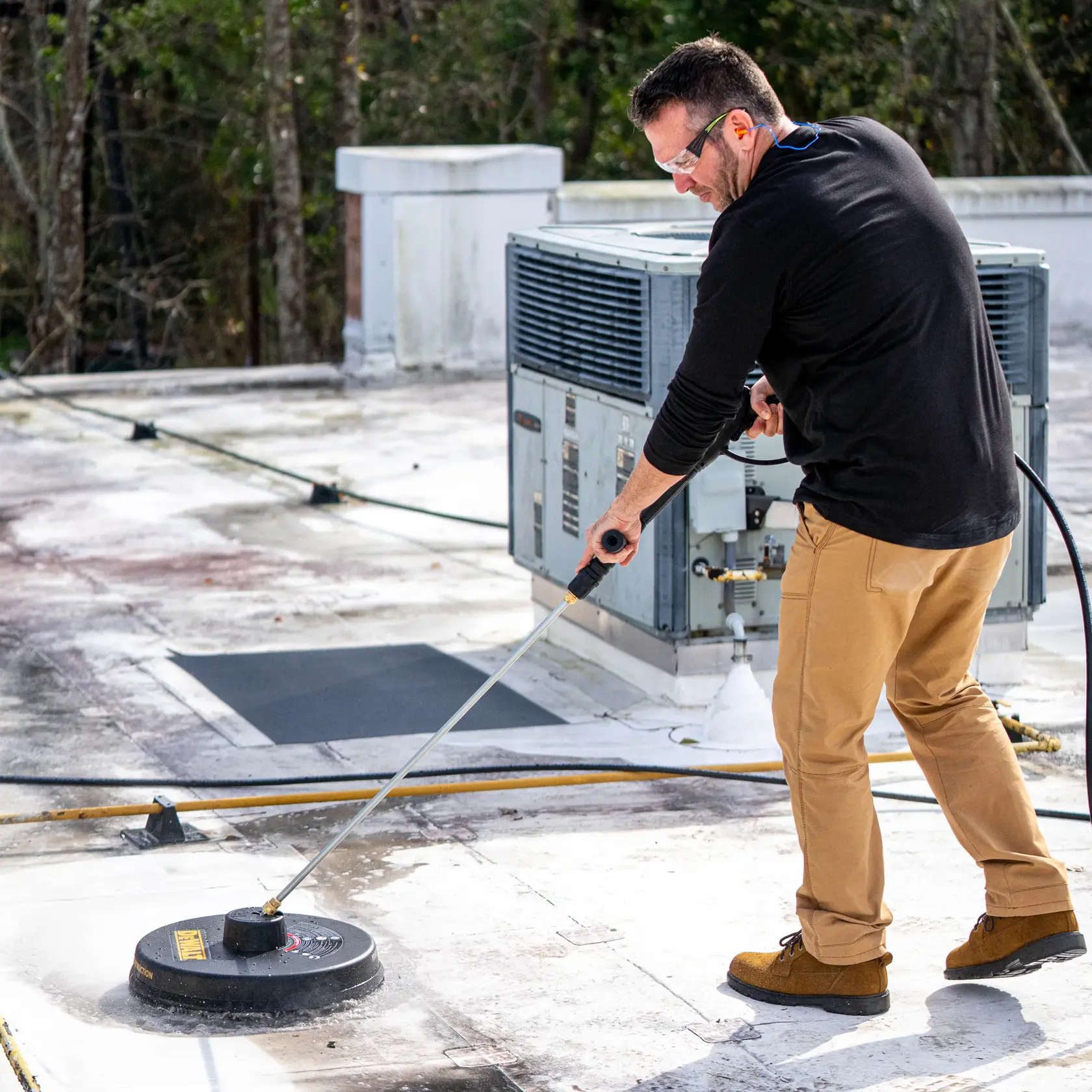A person uses a DEWALT DXPA34SC surface cleaner attachment with a pressure washer to clean a rooftop. The person is standing near an HVAC unit and the surface cleaner is in use on a flat, dirty roof area.