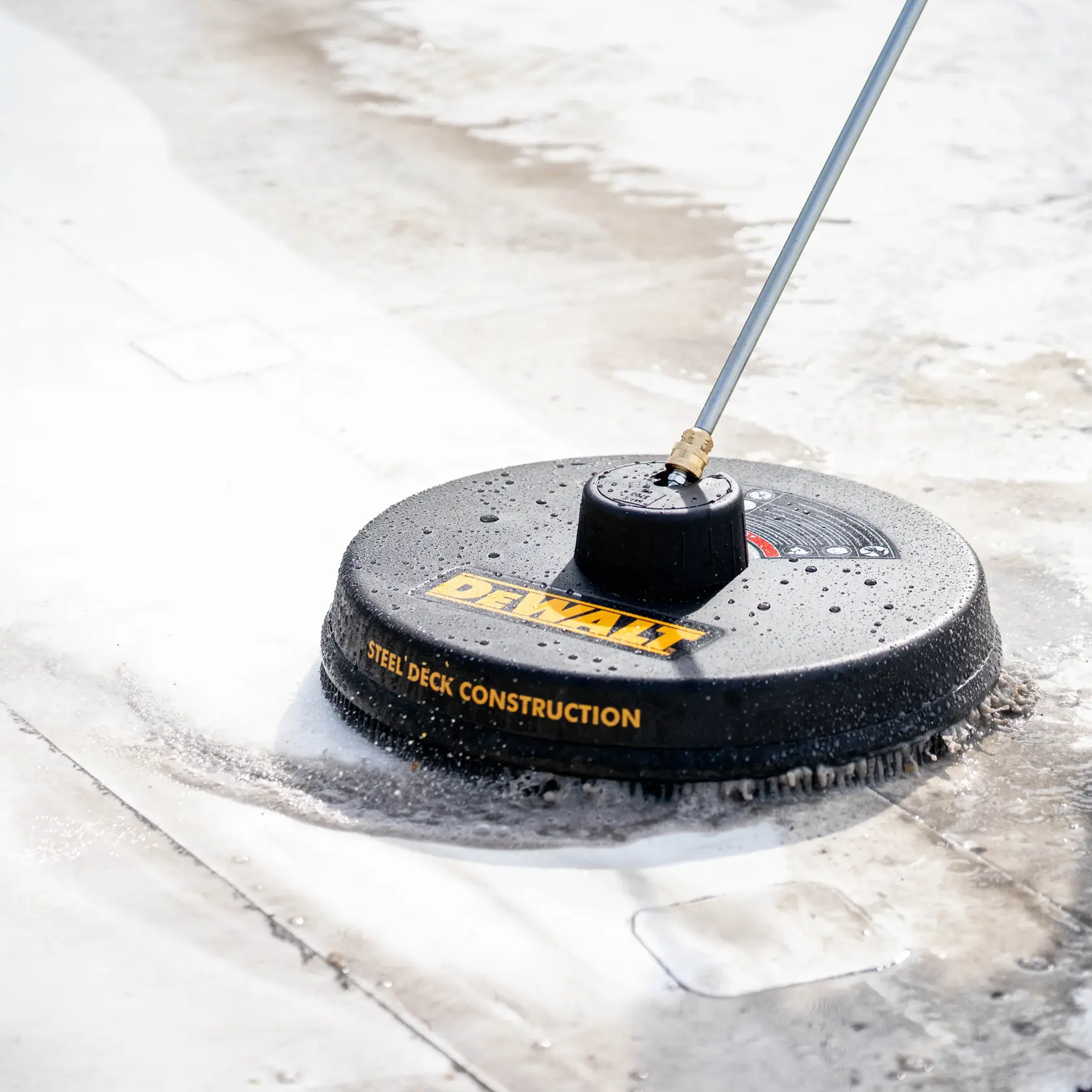A DEWALT steel deck construction surface cleaner is being used to clean a flat outdoor concrete surface, with water and foam visible around the device.