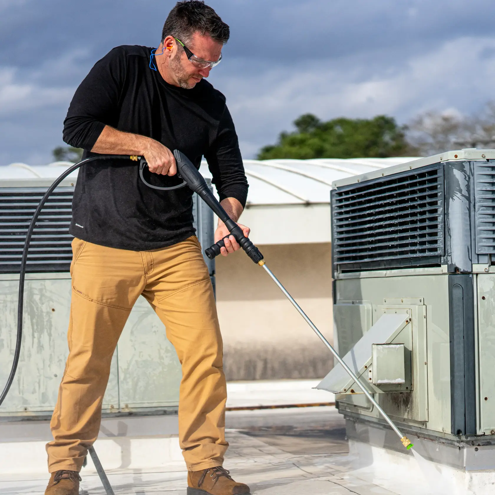 A person using a DEWALT pressure washer to clean the base of an outdoor air conditioning unit on a rooftop, wearing protective ear plugs, khaki pants, and brown shoes.