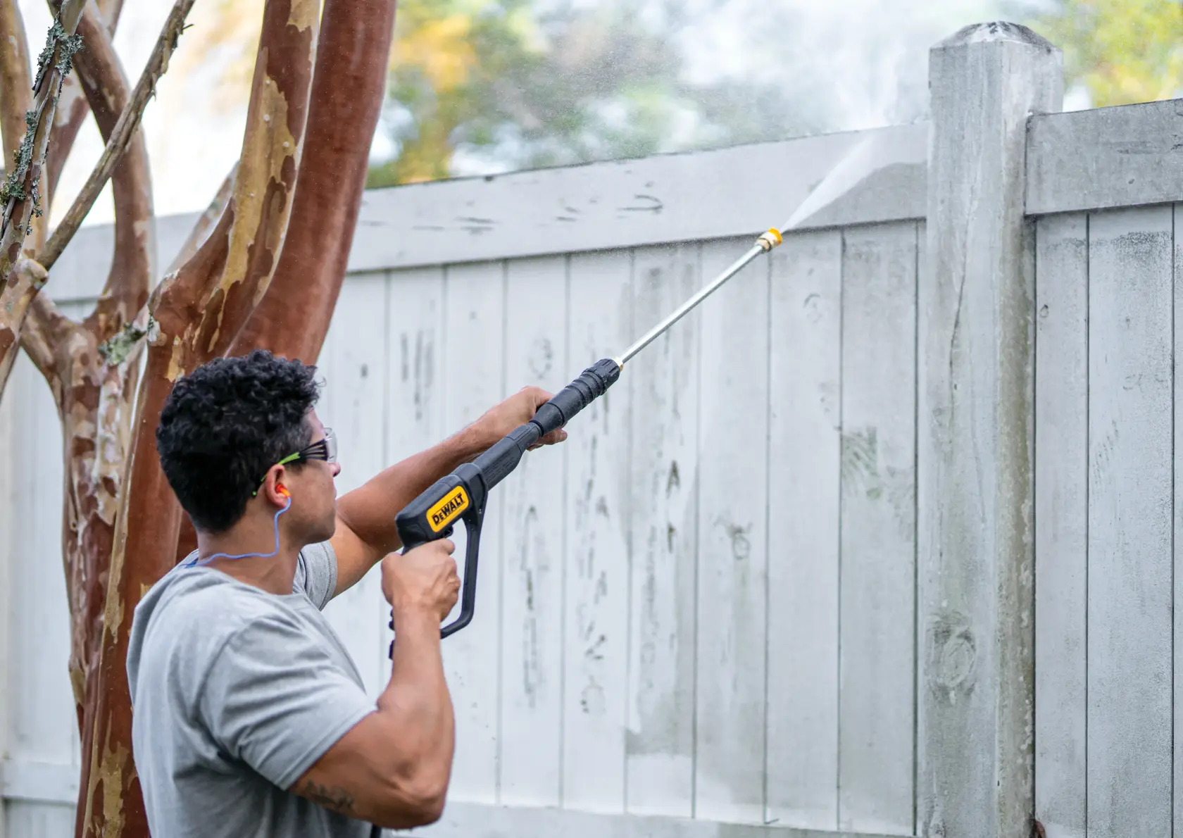 A person using a DEWALT pressure washer to clean a wooden fence outdoors. The individual is holding the washer with both hands and spraying water onto the fence, which has visible dirt and stains.