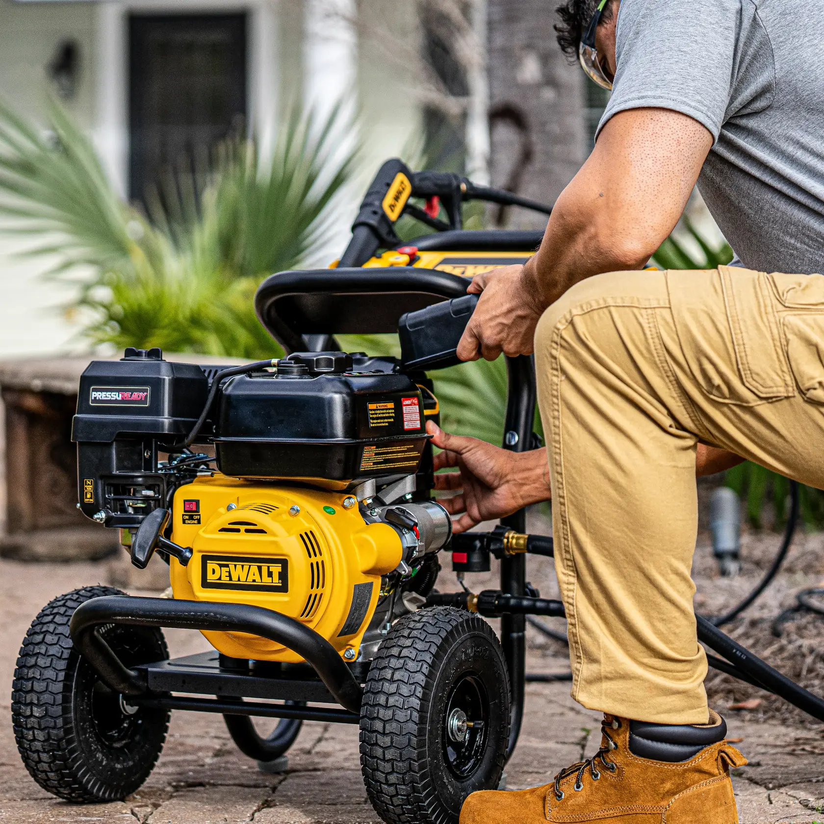 A person in safety goggles and work boots preparing a DEWALT pressure washer outdoors on a paved surface, with green plants and a house in the background.