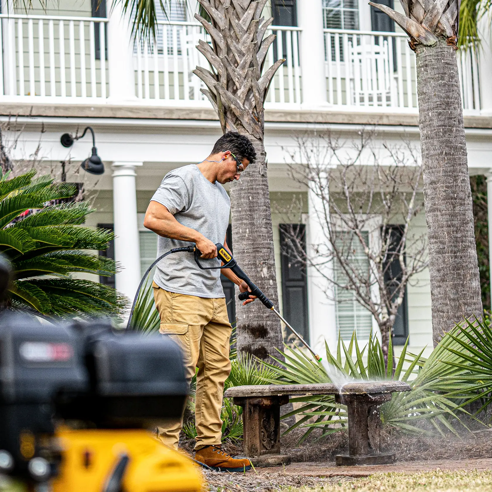 A person using a DEWALT pressure washer to clean an outdoor stone bench in a garden area, with palm trees and a house in the background.