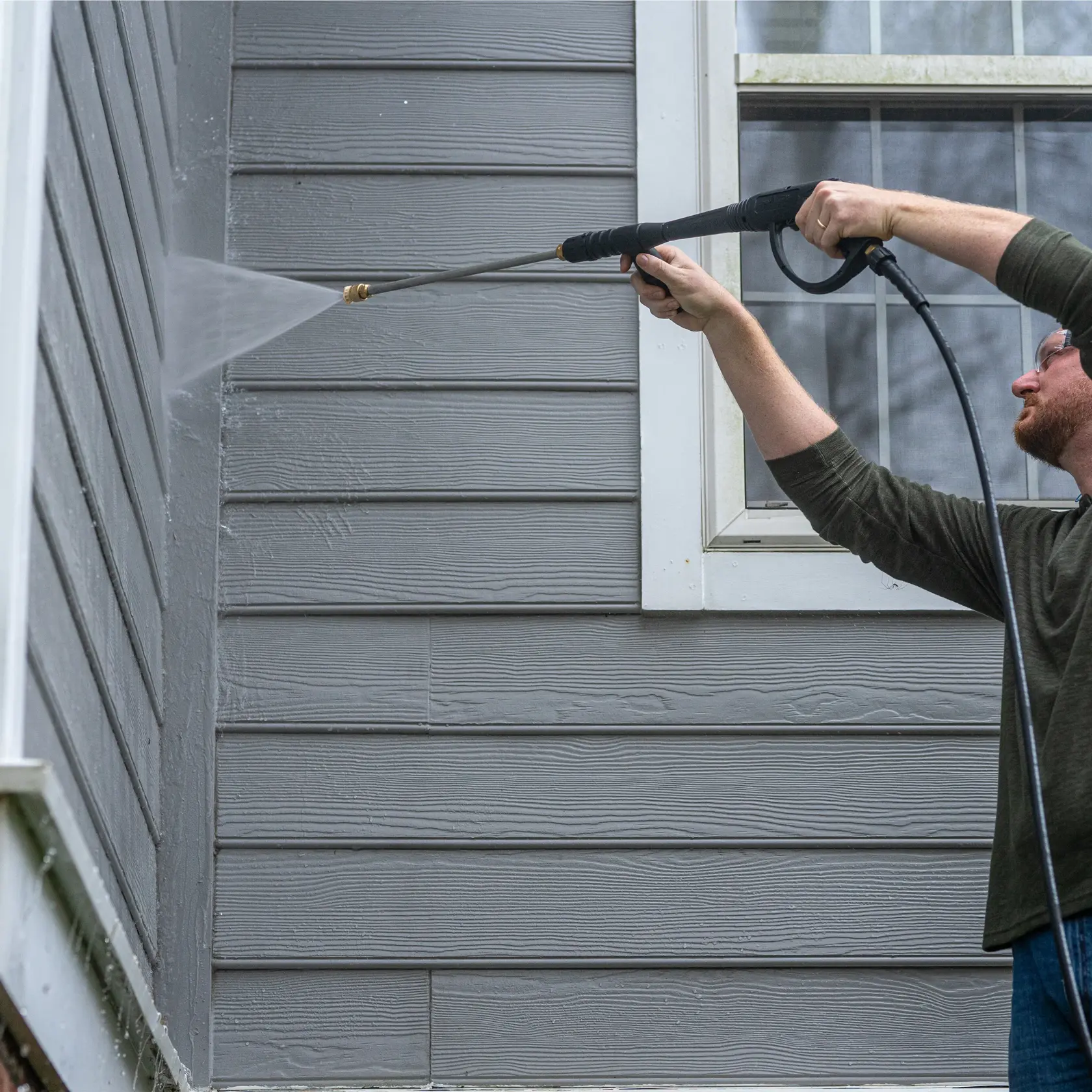 A person using a DEWALT power washer to clean the exterior wall of a house. The water spray is directed at grey siding near a window.