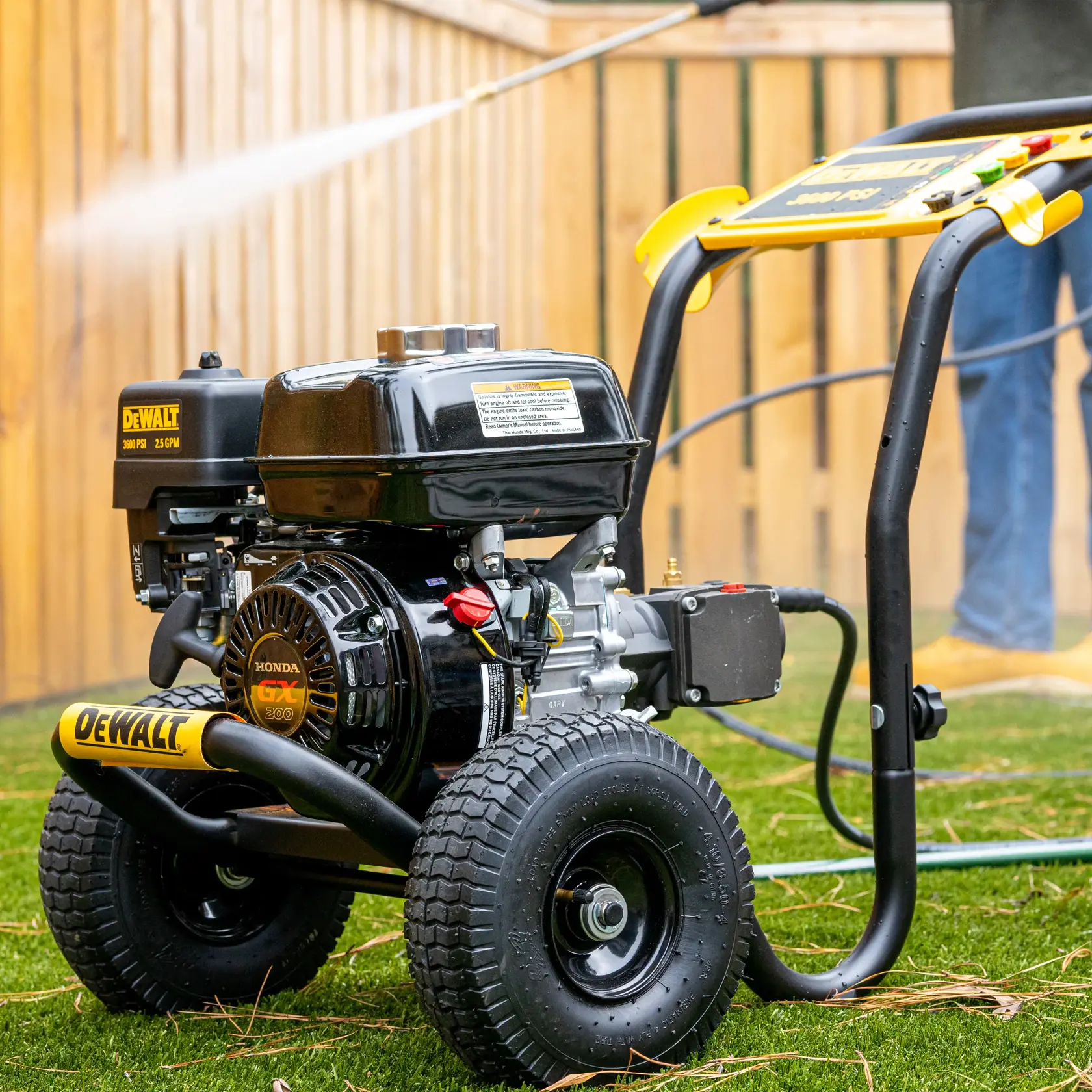 A DEWALT DXPW3625 pressure washer with Honda GX engine is shown on a grassy lawn, spraying water while being operated by a person in the background near a wooden fence.