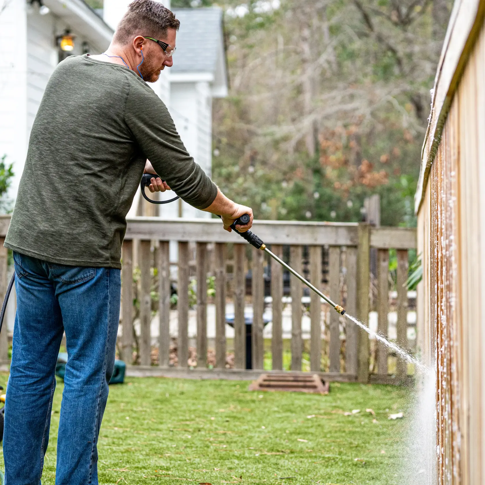 A person is using a DEWALT pressure washer to clean a wooden fence outdoors in a backyard. Water sprays from the washer, removing dirt and debris from the fence.