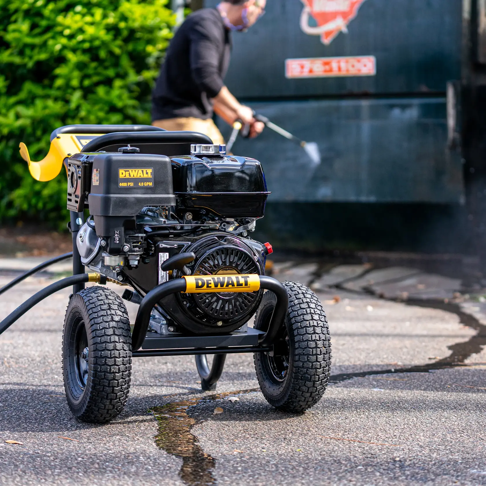 DEWALT DXPW4440 pressure washer on wheels is shown outdoors on pavement, with a person in the background using a spray wand to clean a large surface.