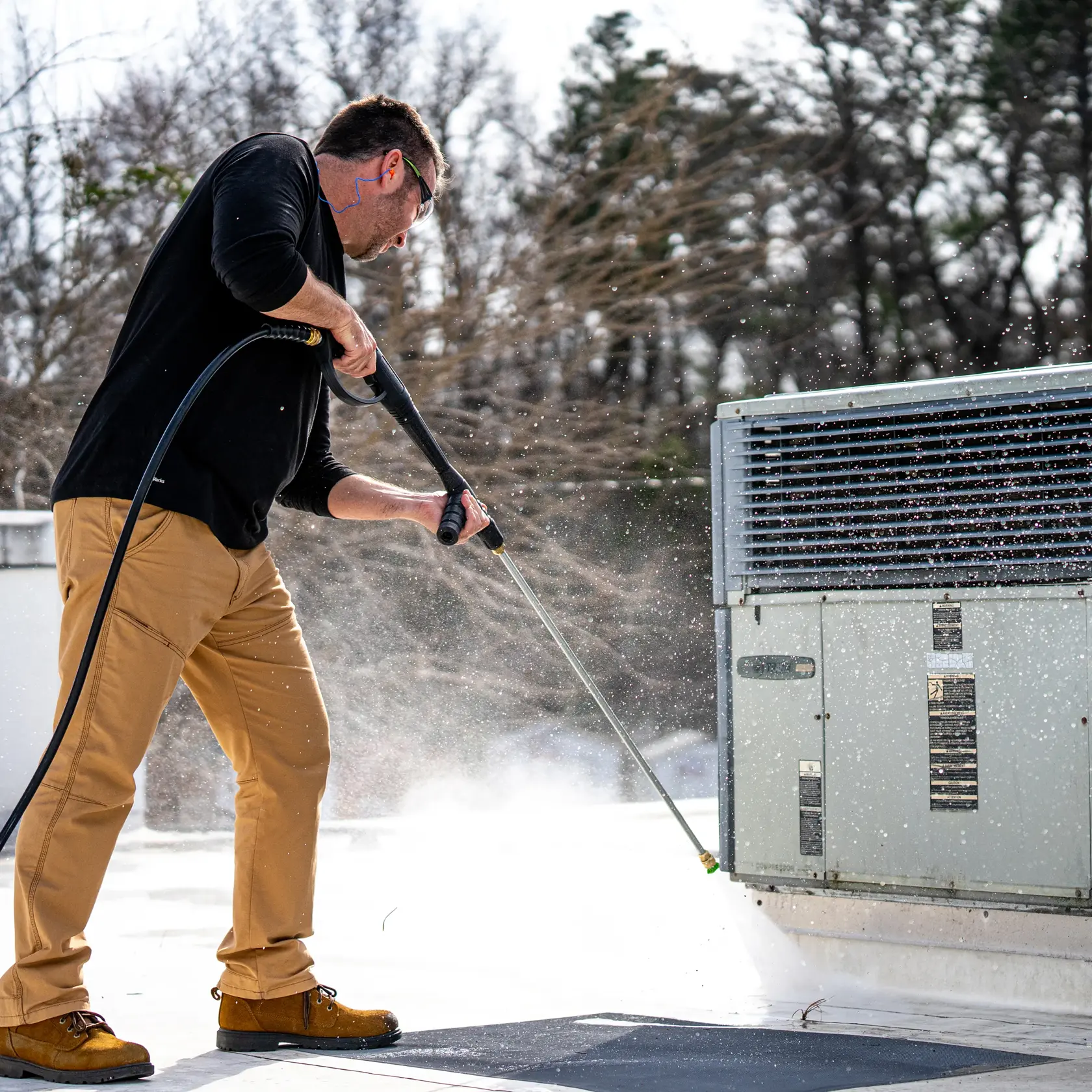 A person wearing tan pants and brown work boots is using a DEWALT pressure washer to clean an outdoor industrial unit on a rooftop. Water sprays around as the person directs the washer at the unit. Trees are visible in the background.