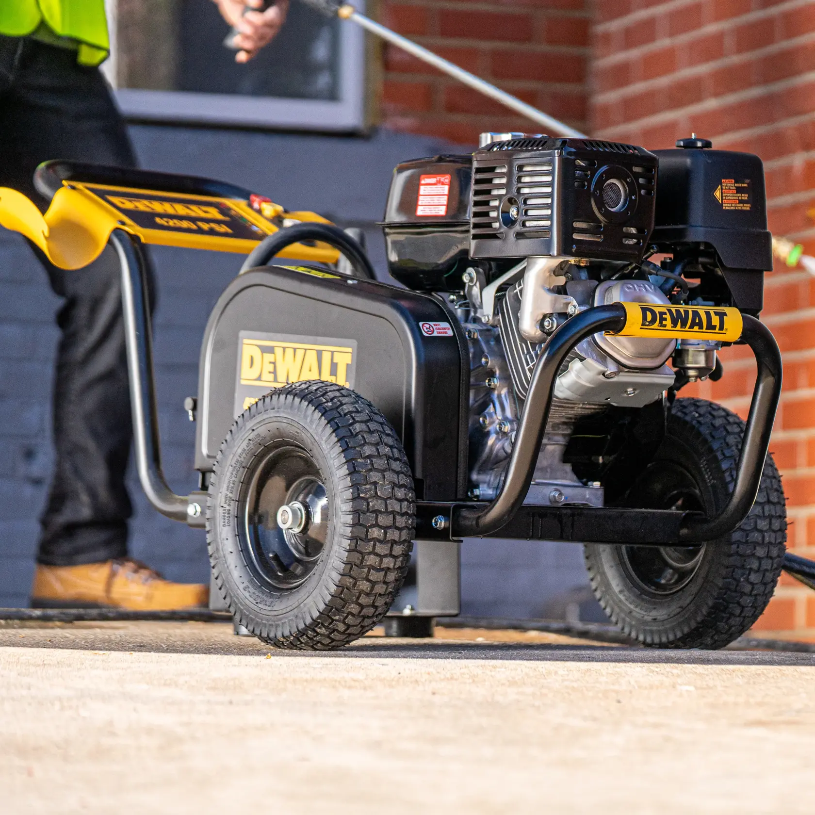 A DEWALT DXPW60606 pressure washer with sturdy wheels is shown outdoors, with a person using it to clean a surface. The machine is black and yellow in color and placed on a concrete area near a brick wall.