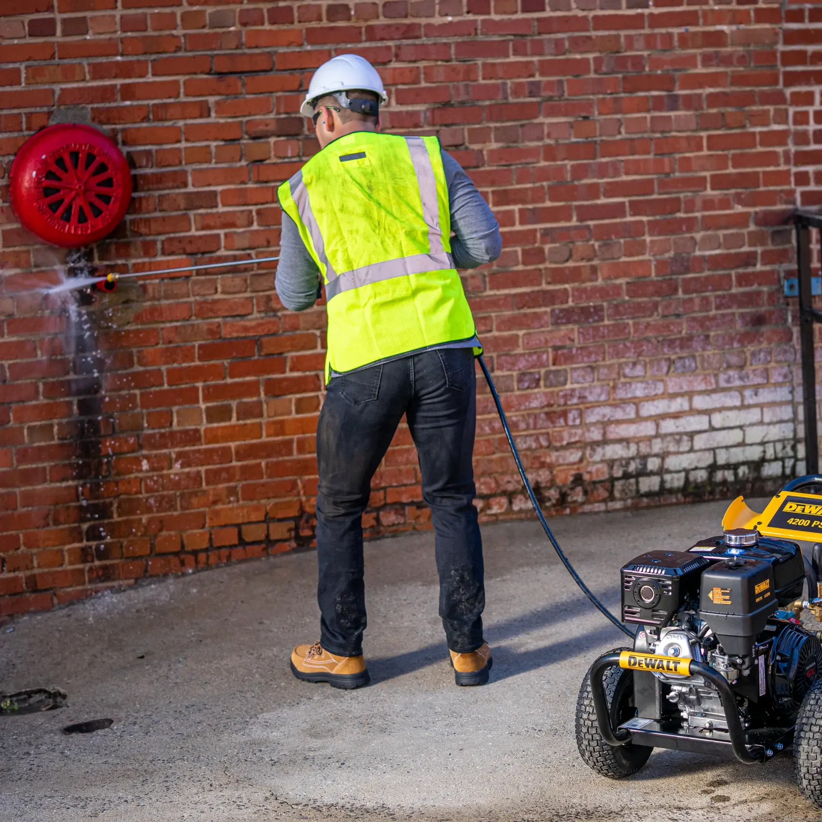A person wearing a hard hat and a reflective safety vest is using a DEWALT pressure washer to clean a brick wall. The DEWALT pressure washer is visible on the right side of the image.